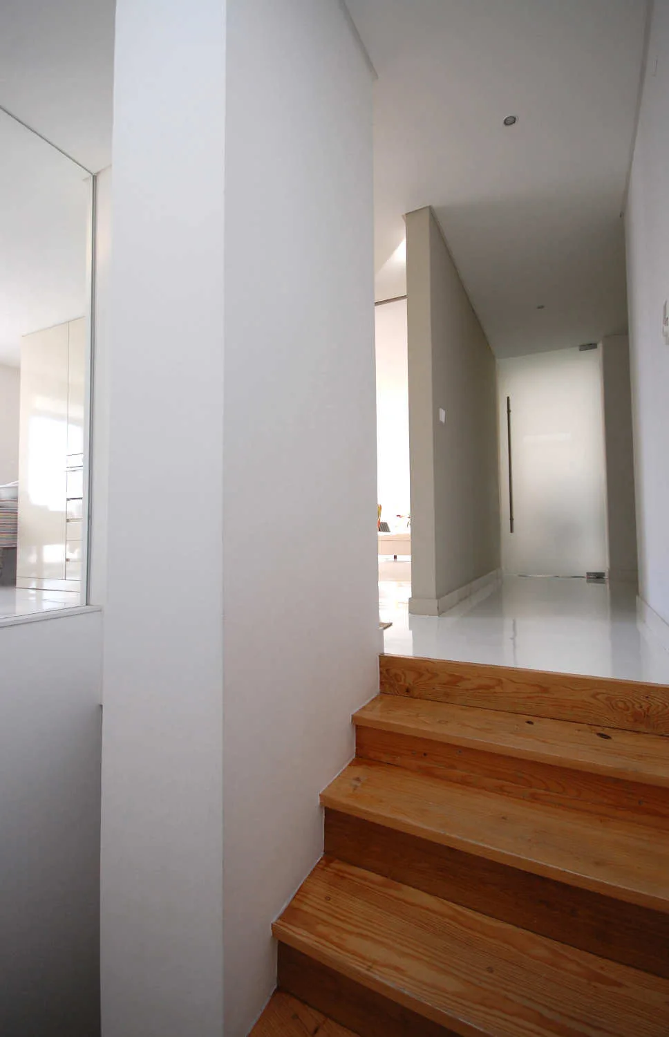 A wooden staircase leads up to a bright white minimalist hallway with a glossy floor and a frosted glass door at the end.