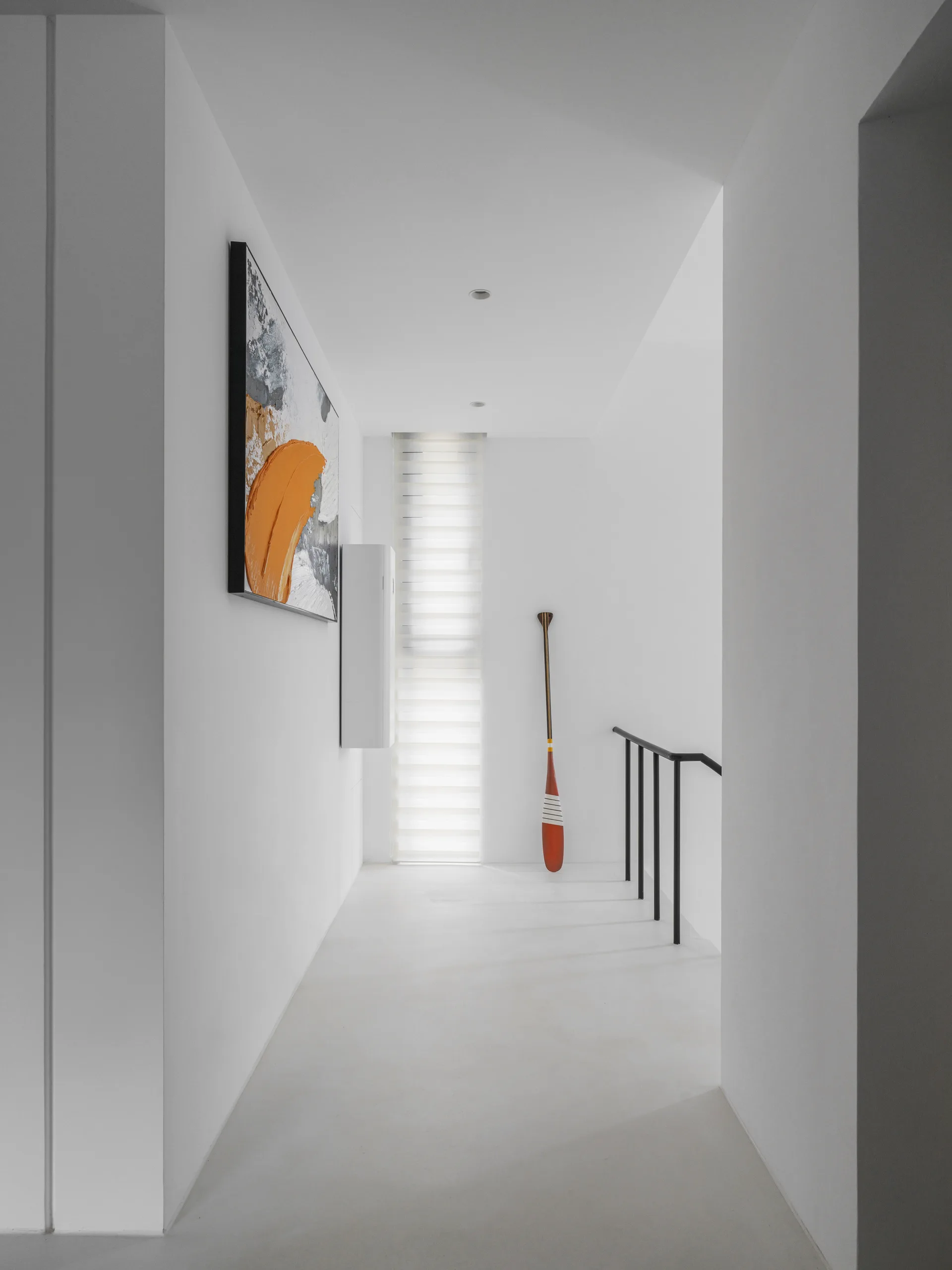 A minimalist white hallway featuring a narrow window with blinds, a black metal staircase railing, an abstract painting, and a decorative wooden oar.