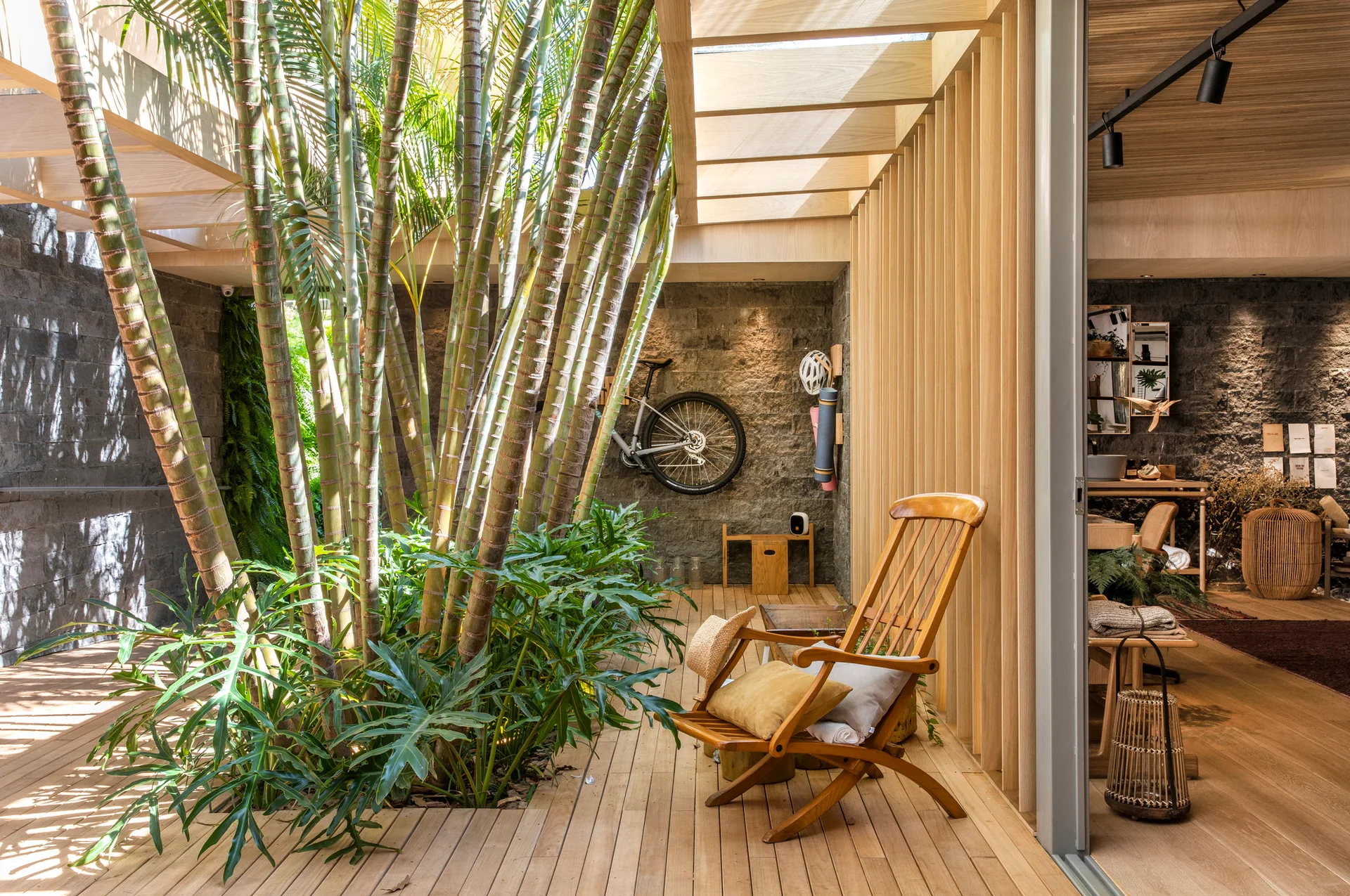 An indoor-outdoor patio with wooden decking features tall palm trees growing through an open ceiling next to a textured charcoal stone wall.