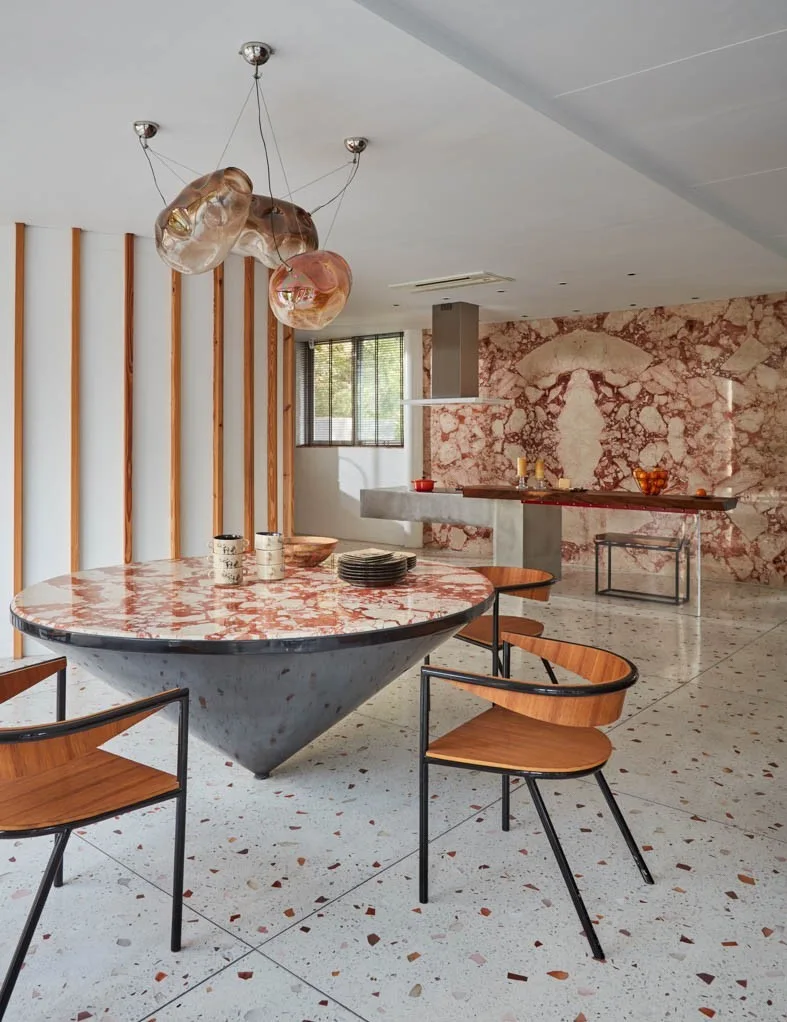 A contemporary dining room features a round red marble table on a conical base, wood and metal chairs, terrazzo flooring, and a matching marble wall.