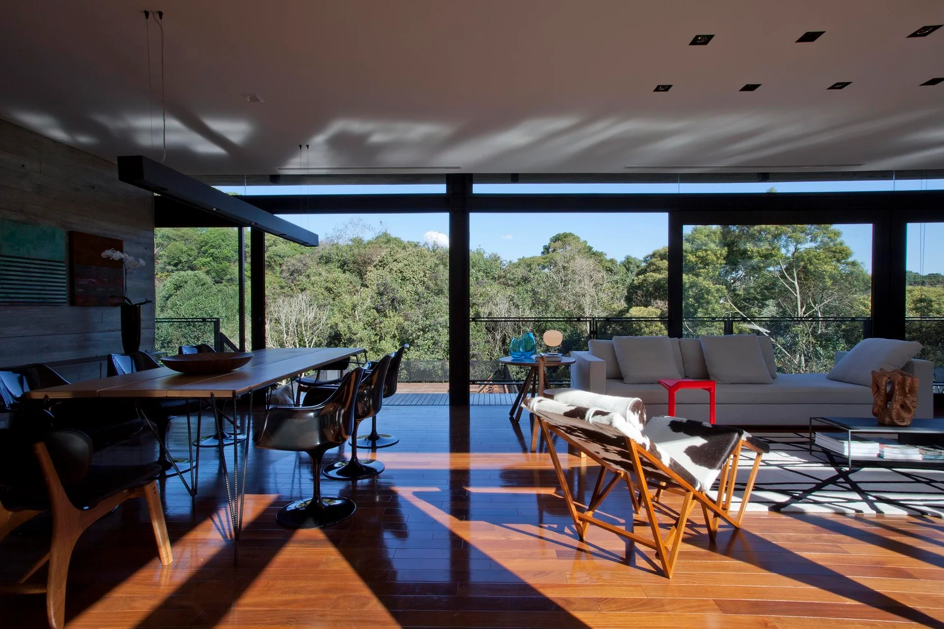 Modern open-concept living room featuring polished wood floors, a light sofa, mid-century dining table, and large windows looking out at a forest.