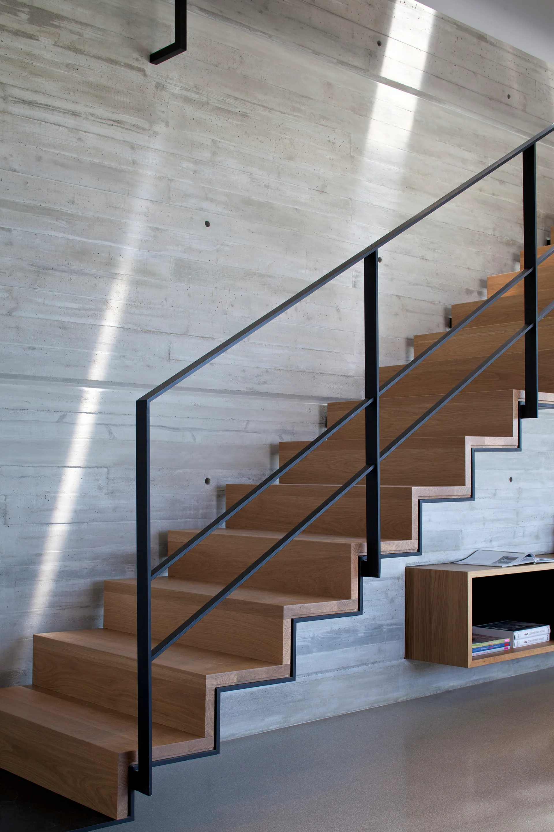 A modern floating wooden staircase with a minimalist black steel handrail rises against a textured board-formed concrete wall.