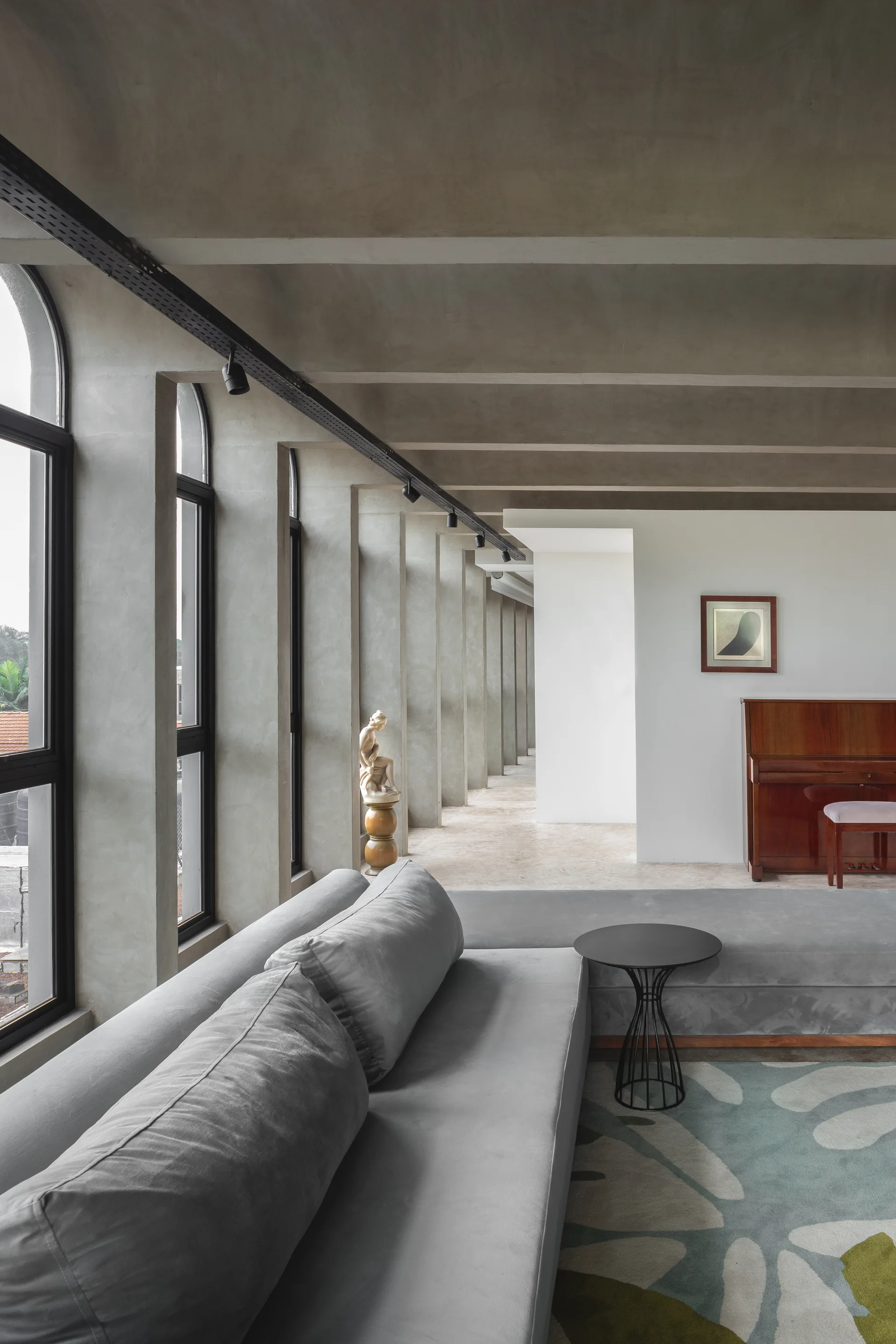A long minimalist living room features exposed concrete ceiling beams, a row of tall arched windows, a gray sofa, and a wooden upright piano.