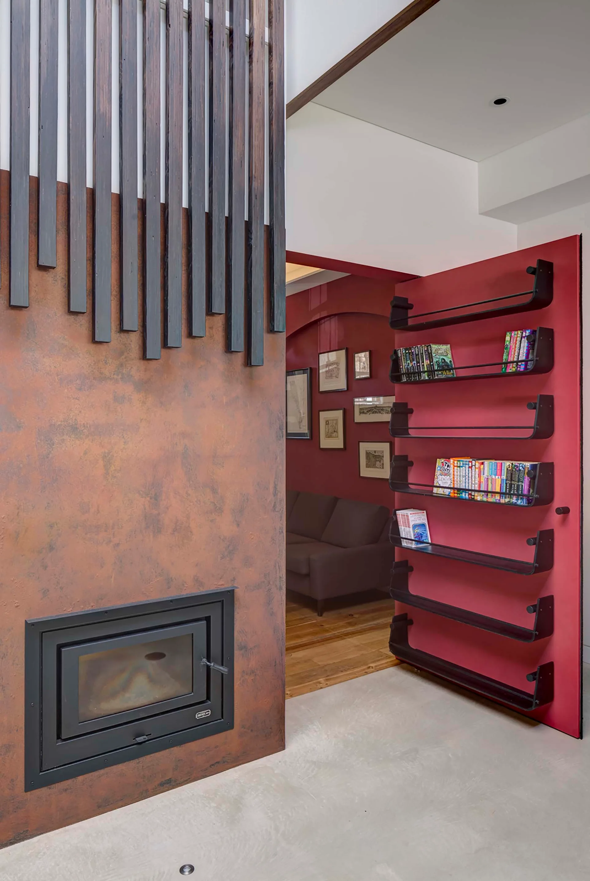 A textured rust-colored fireplace wall with black vertical slats stands next to a vibrant red door featuring black metal book ledges.