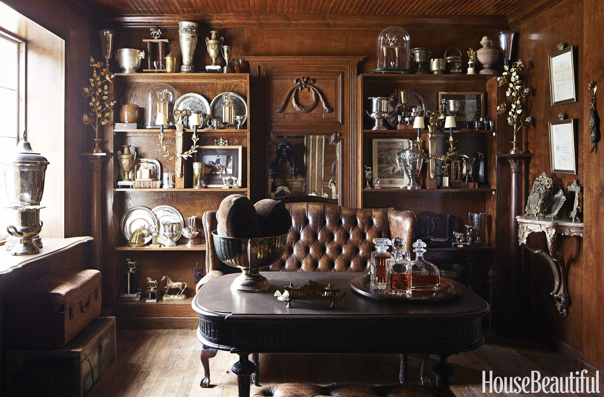 A dark wood-paneled study features built-in shelves filled with silver trophies, a tufted brown leather sofa, and a desk with glass decanters.