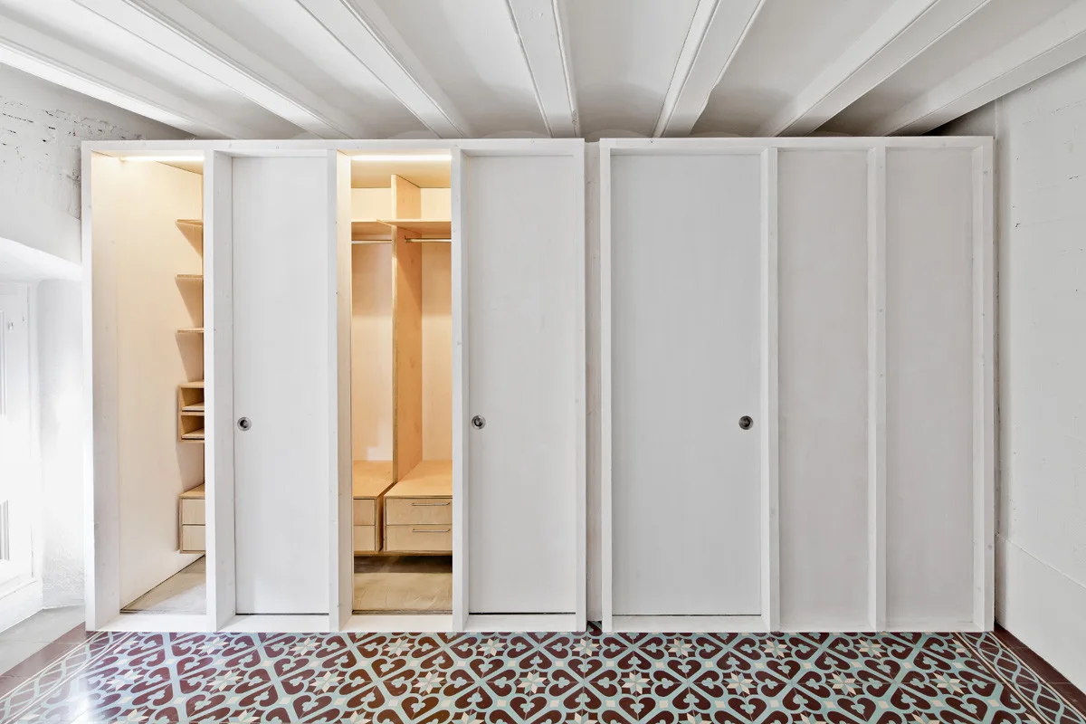 A minimalist white built-in wardrobe with sliding doors open to reveal warm wood shelving and drawers over a patterned tile floor.