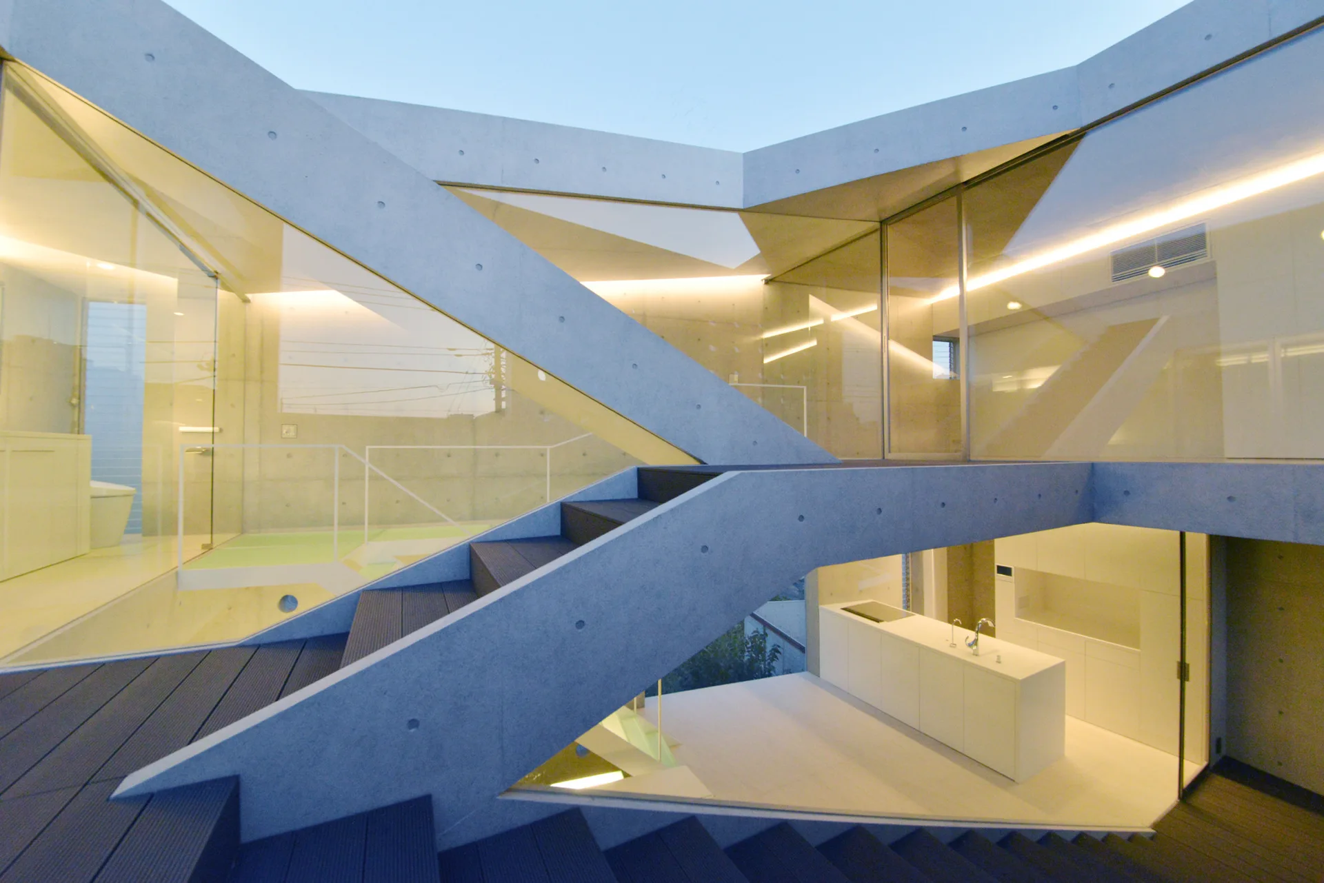 An exposed concrete staircase with glass railings winds through a minimalist multi-level home, overlooking a white kitchen island below.