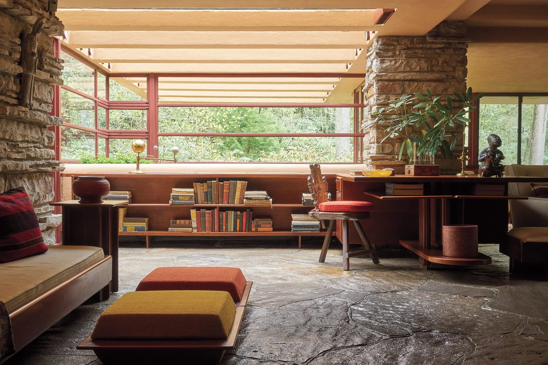 A rustic living room features flagstone flooring, stacked stone walls, a low-profile wooden bookcase, and windows with red framing.