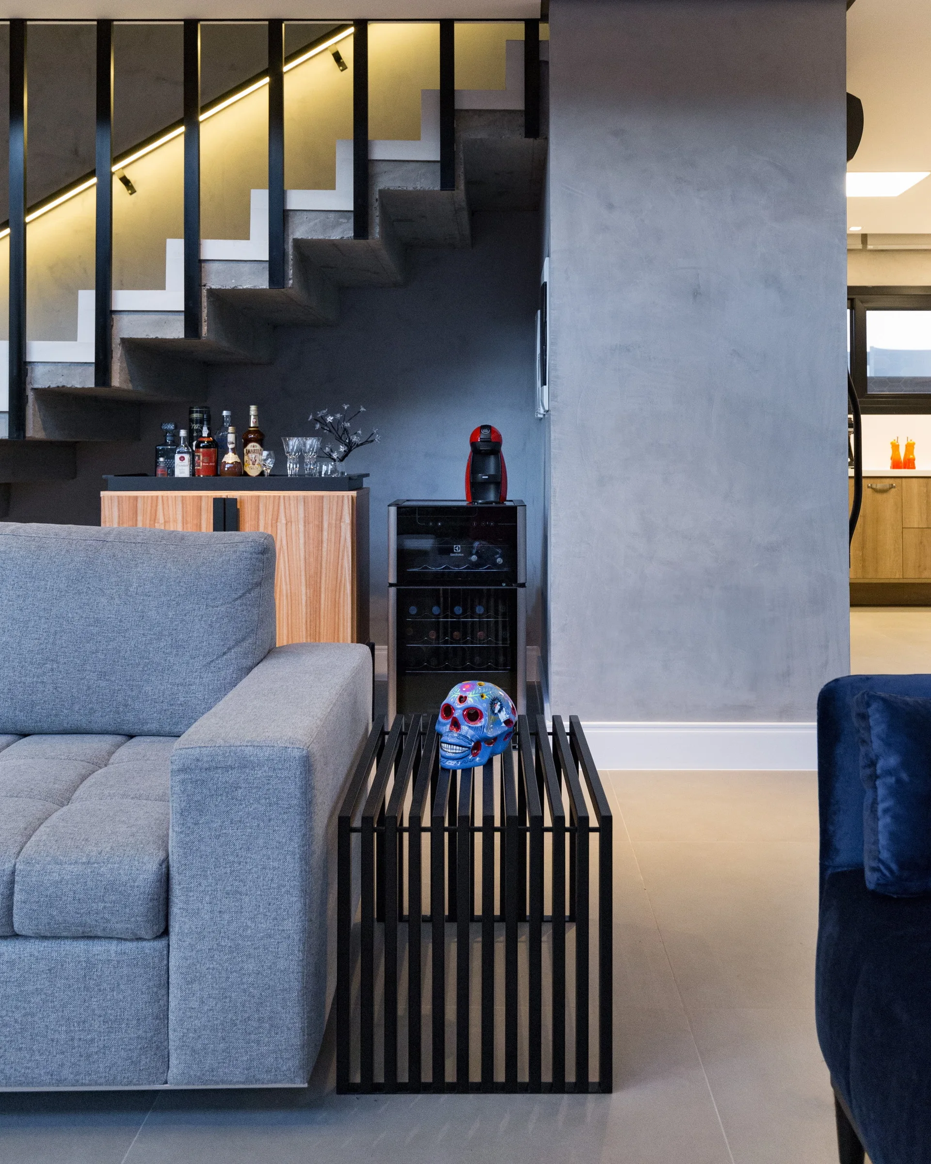 A modern living room features a gray sofa, a slatted black table, and a home bar built into the space beneath a concrete staircase.