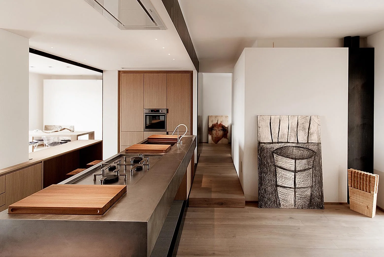 A minimalist kitchen features a long metal island with a built-in cooktop, light wood cabinets, and large abstract artwork leaning against a white wall.