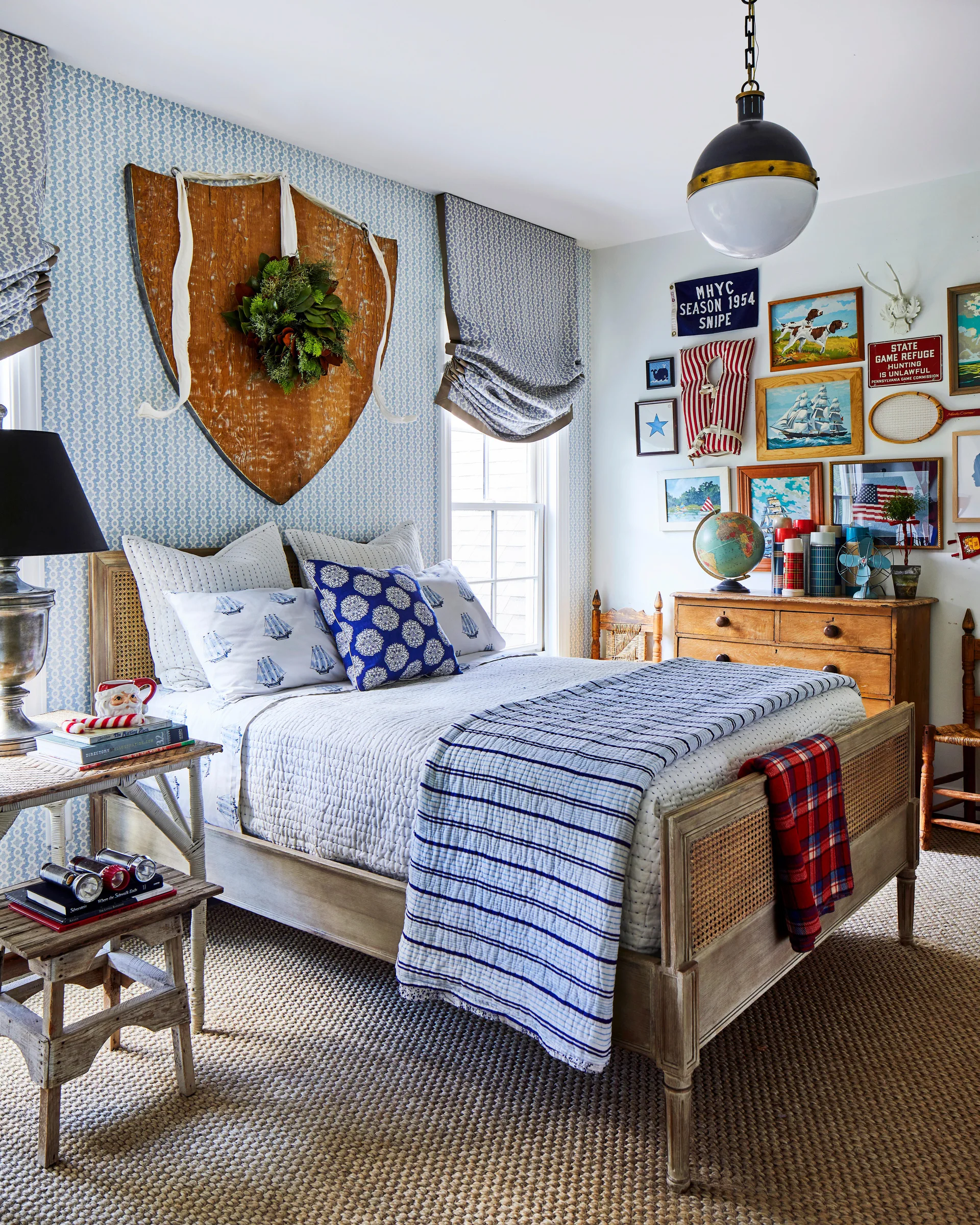 A bedroom features a cane wood bed frame, a large wooden shield wall decoration with a wreath, blue patterned wallpaper, and a vintage gallery wall.