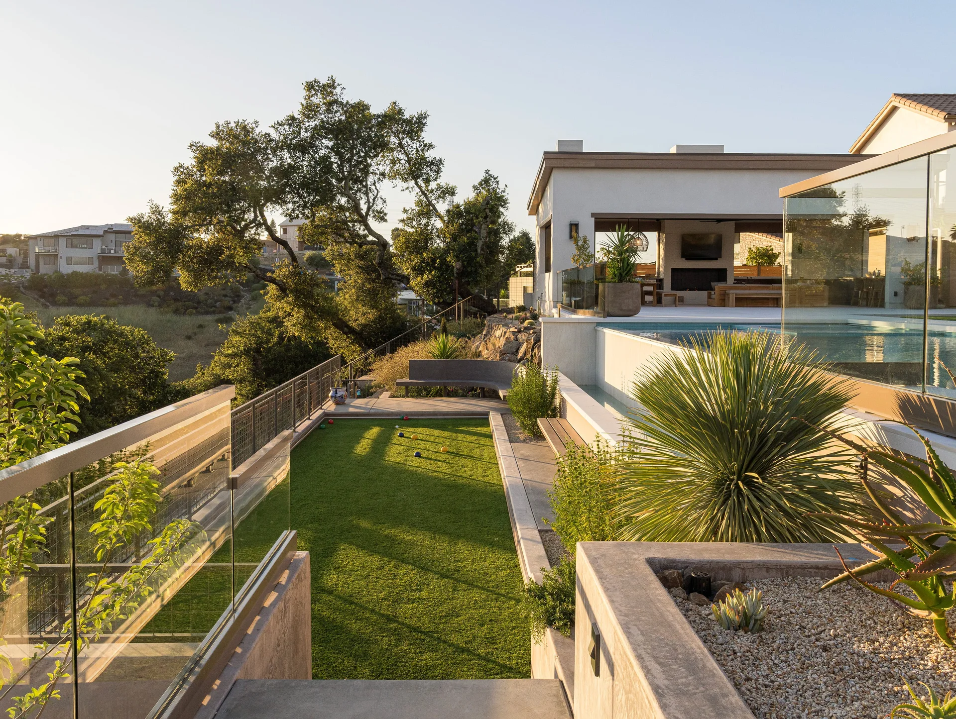 A multi-level modern backyard featuring a rectangular swimming pool with glass railings, a green bocce court, and concrete steps surrounded by plants.