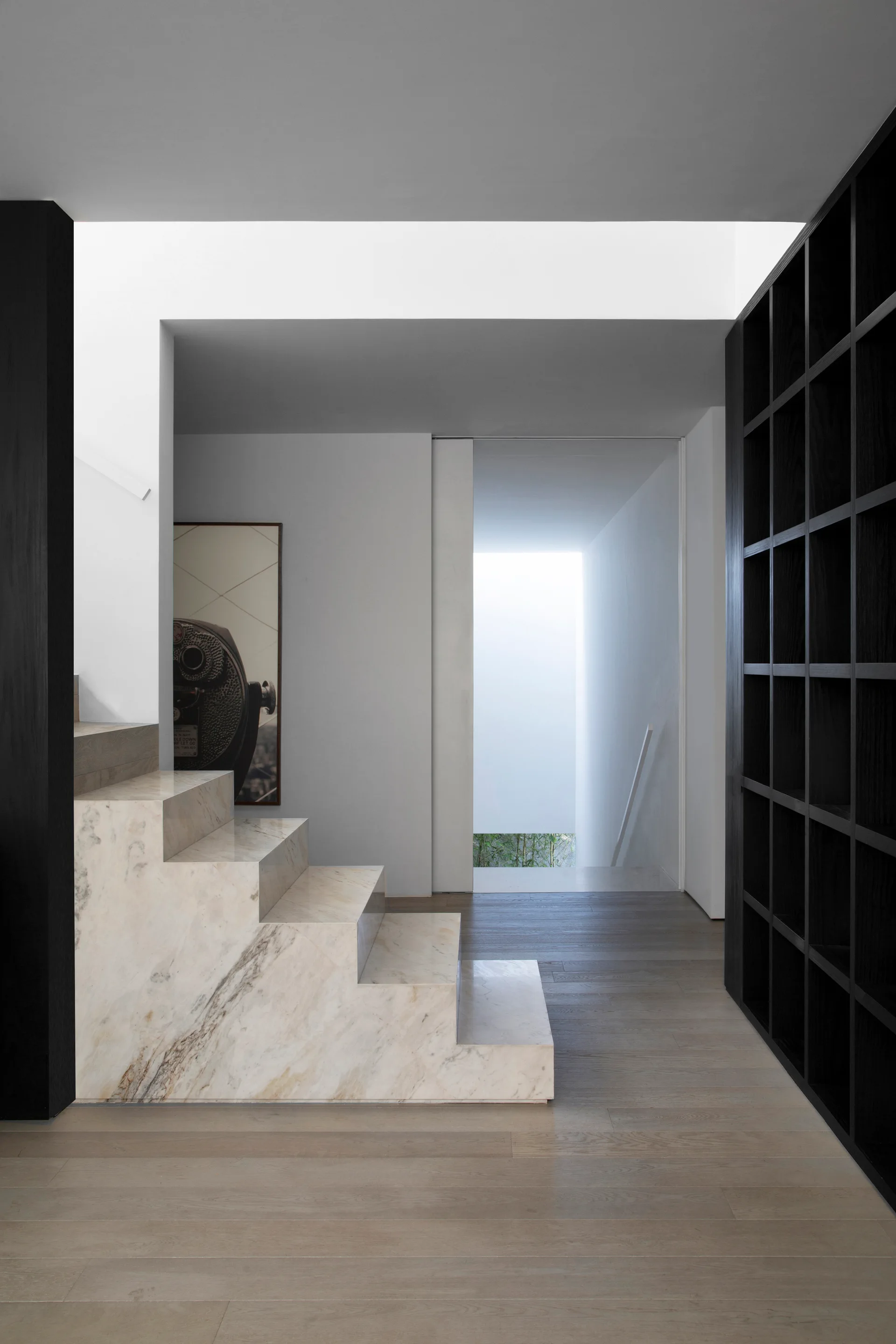 A minimalist foyer features a veined marble staircase next to light wood floors and a tall black grid shelving unit.