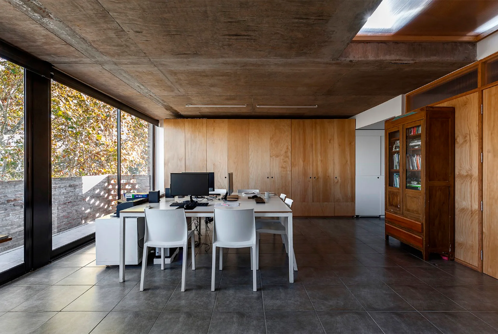 A minimalist home office features a large central desk with white chairs, a raw concrete ceiling, and a full wall of wood cabinetry.