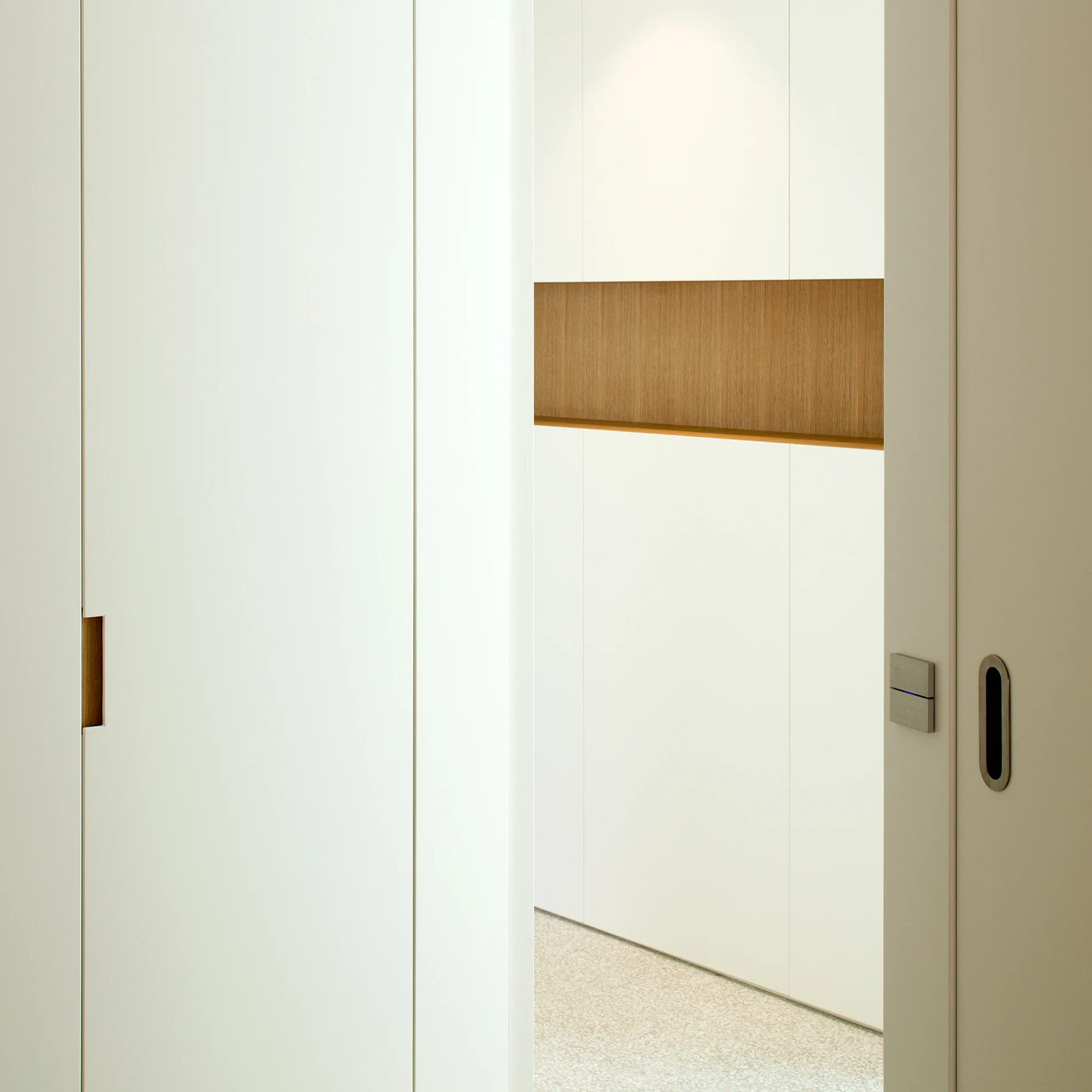 A minimalist white door with a recessed wood handle opens to a hallway with terrazzo flooring and a horizontal wood-toned wall recess.