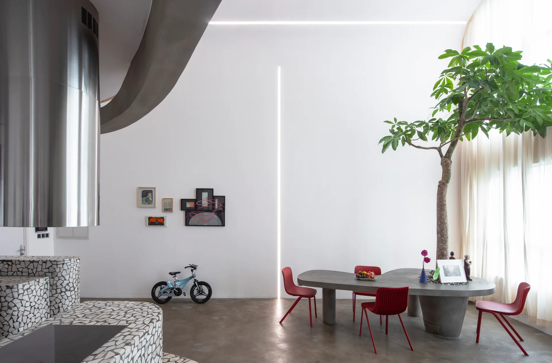 A minimalist dining room with a concrete-style table holding an integrated living tree, paired with red chairs and a mosaic-tiled kitchen counter.