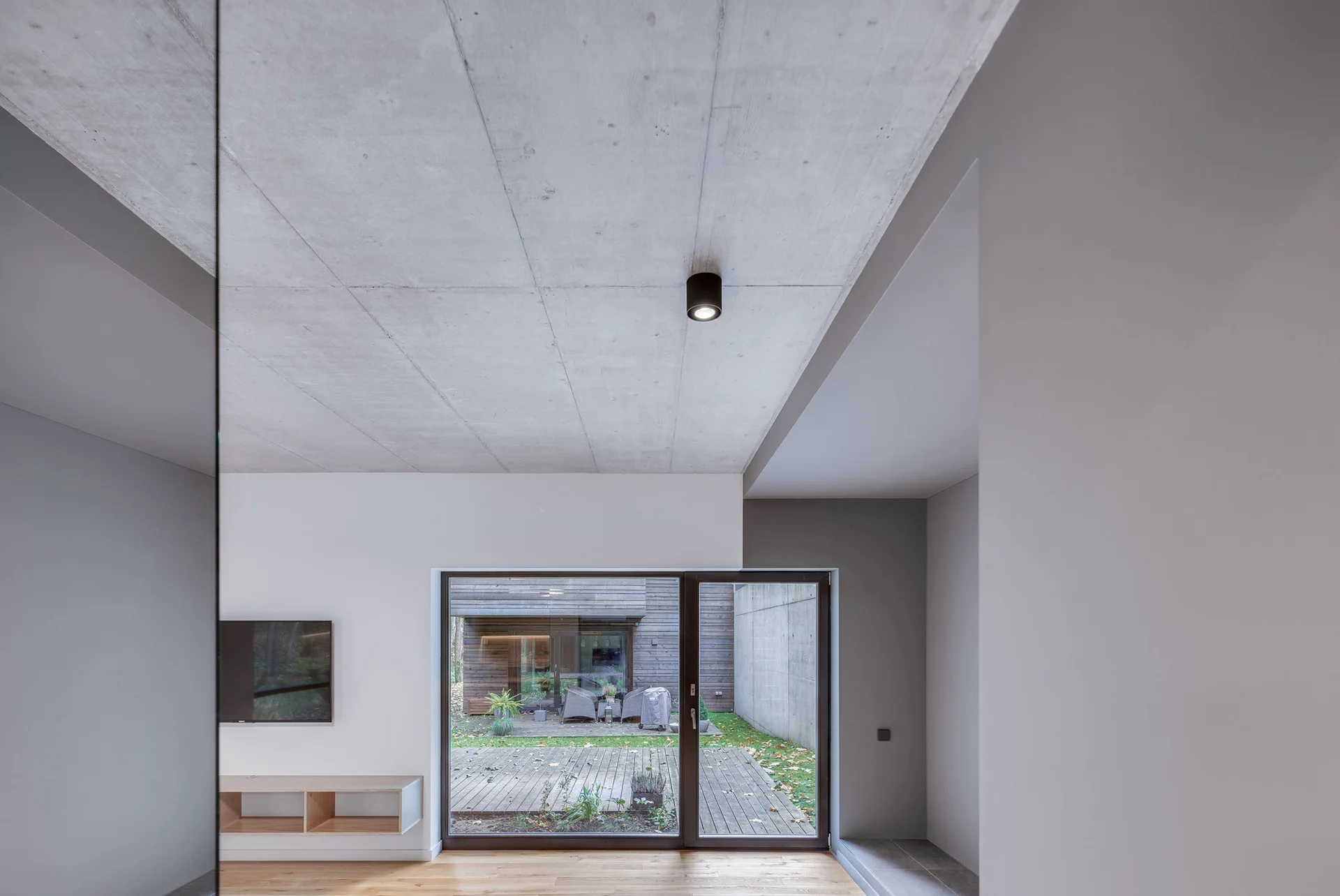 A minimalist room features a textured grey concrete ceiling, white walls, light wood floors, and a sliding glass door looking out to an outdoor deck.