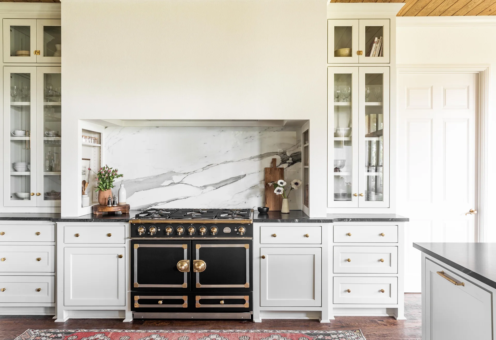 A residential kitchen features off-white cabinetry, a black and brass range, a veined marble backsplash, and symmetrical glass-front upper cabinets.