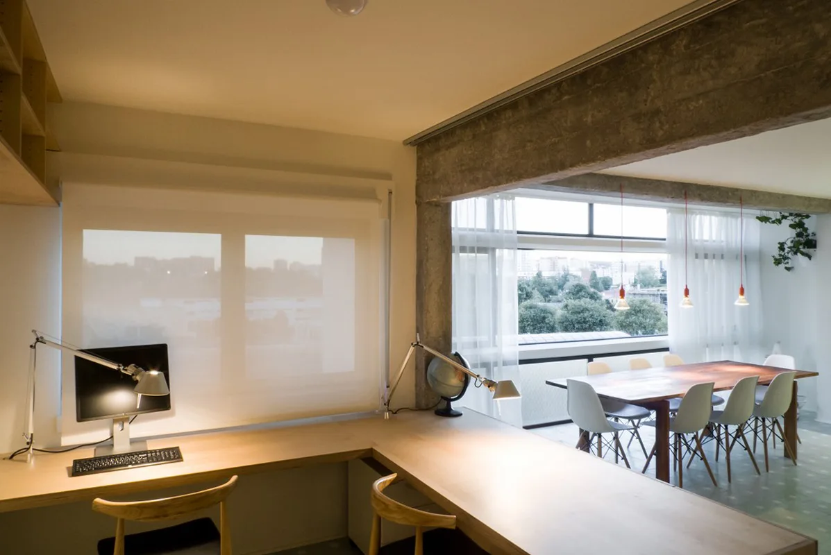 An open-plan room showing a light wood corner desk in the foreground and a dining table beneath an exposed concrete ceiling beam.