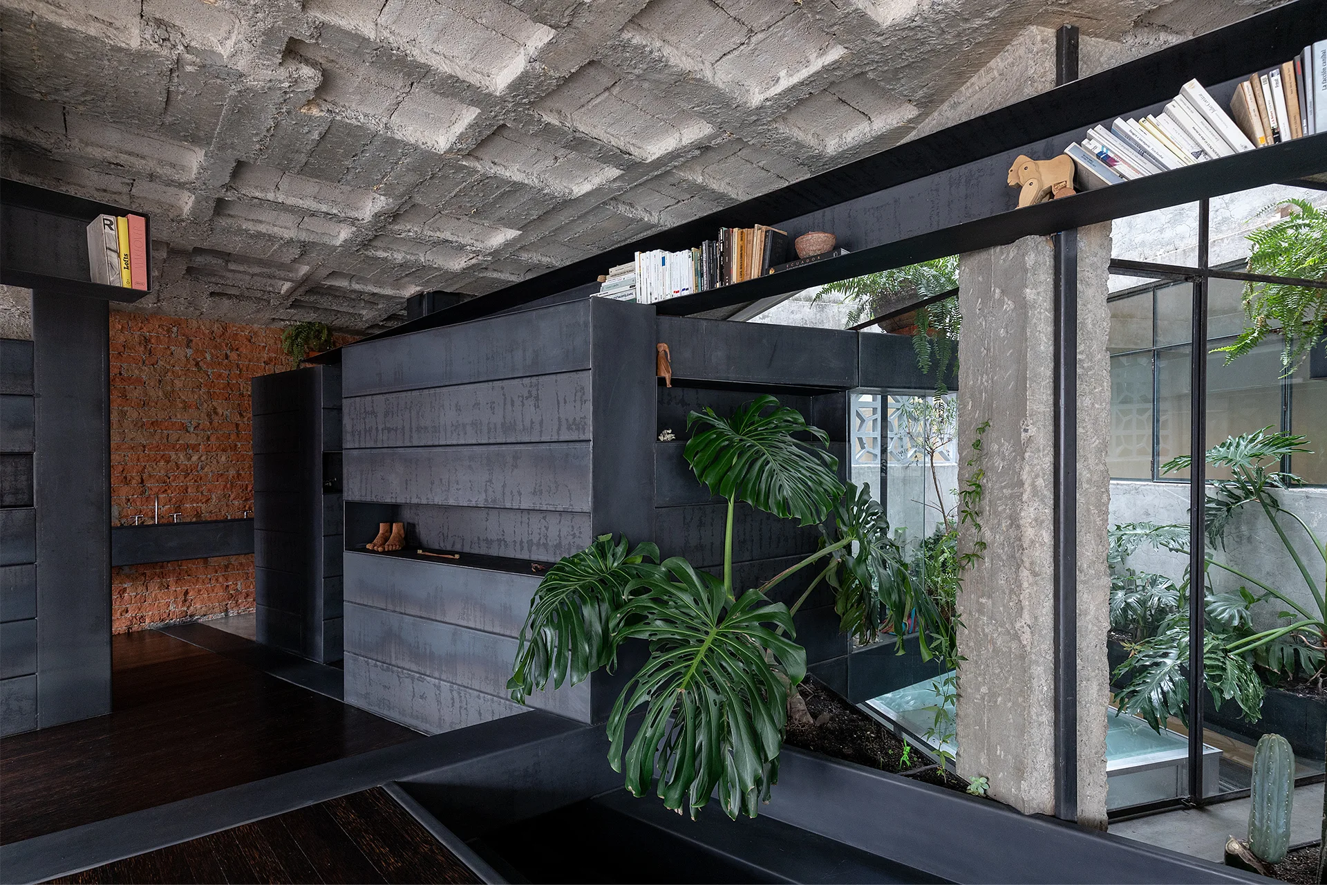 An industrial interior features an exposed concrete waffle ceiling, dark metal modular partitions, and an indoor garden planter with a large monstera.