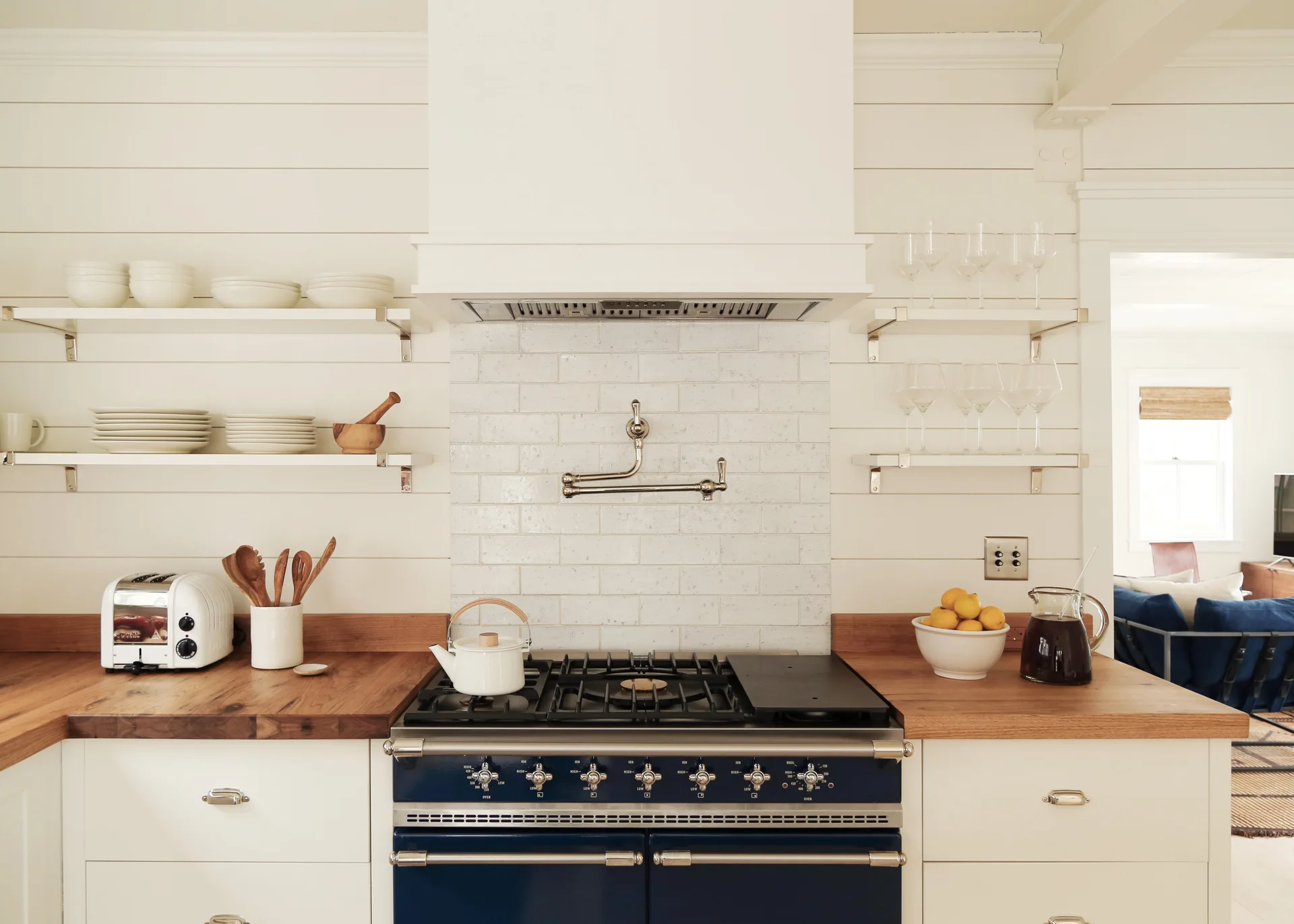 A bright kitchen features a blue gas range under a white hood, surrounded by shiplap walls, open shelving, and warm butcher block countertops.
