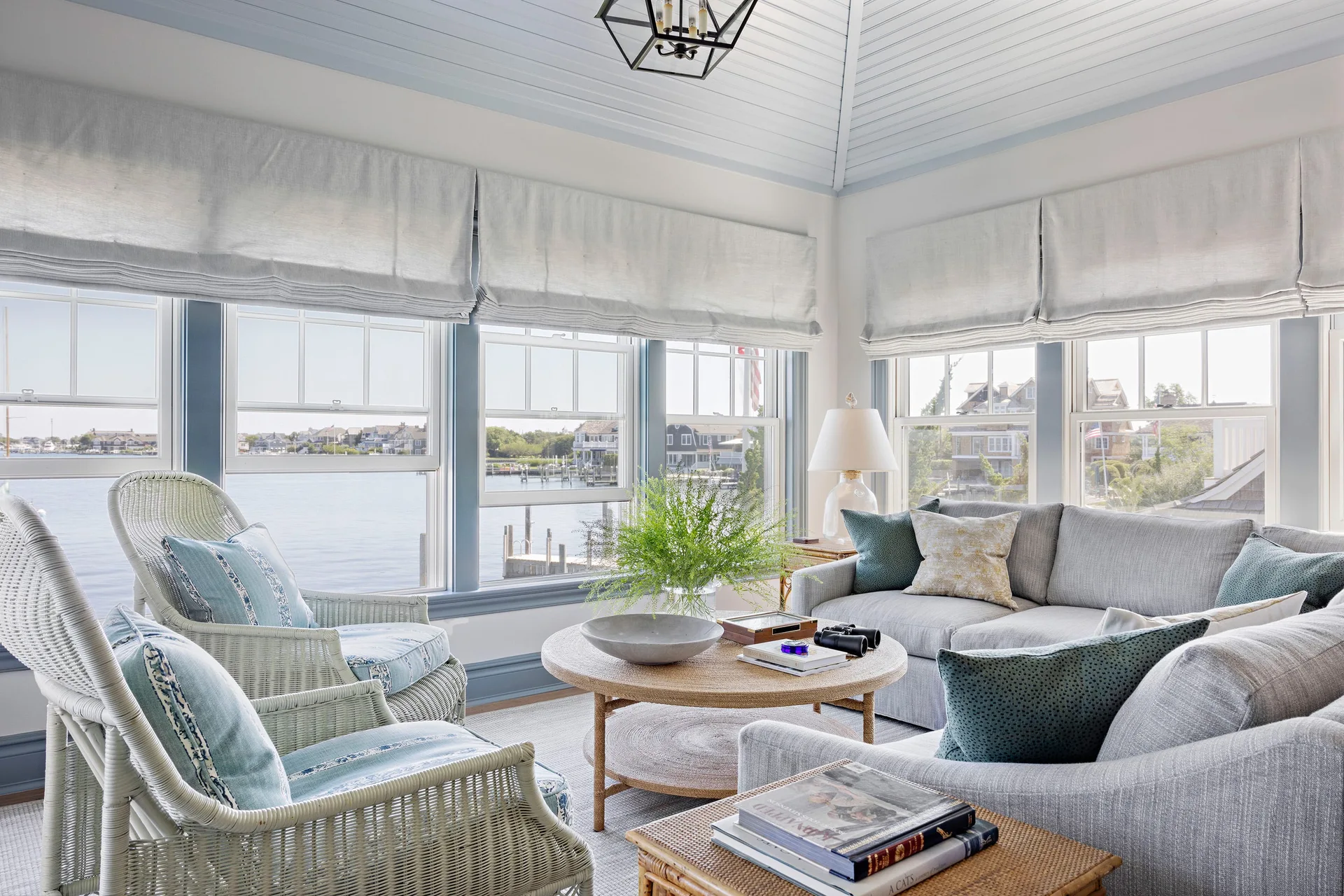 A coastal sunroom features a grey sectional sofa, white wicker armchairs, and a round woven coffee table in front of large windows overlooking water.
