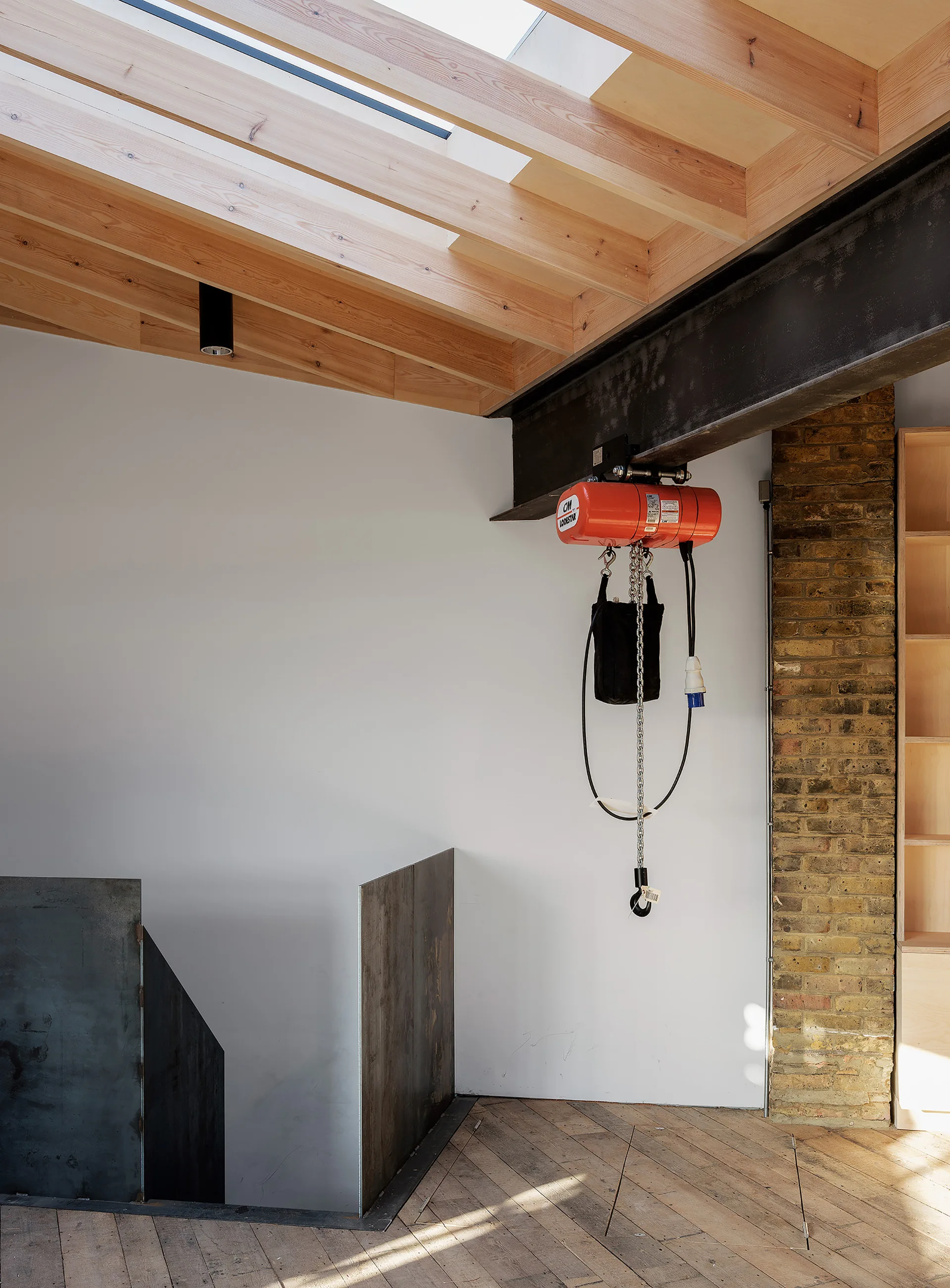 An industrial stairwell features a steel beam with a red electric hoist under an exposed timber ceiling with a skylight.