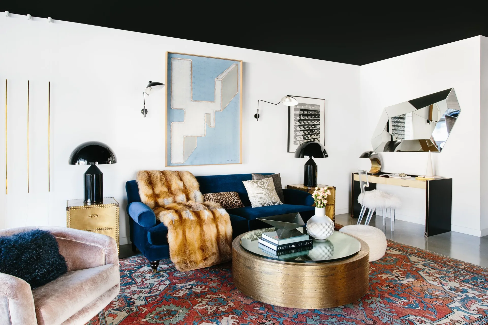 A living room featuring a navy velvet sofa with a fur throw, a black painted ceiling, a brass coffee table, and a patterned area rug.