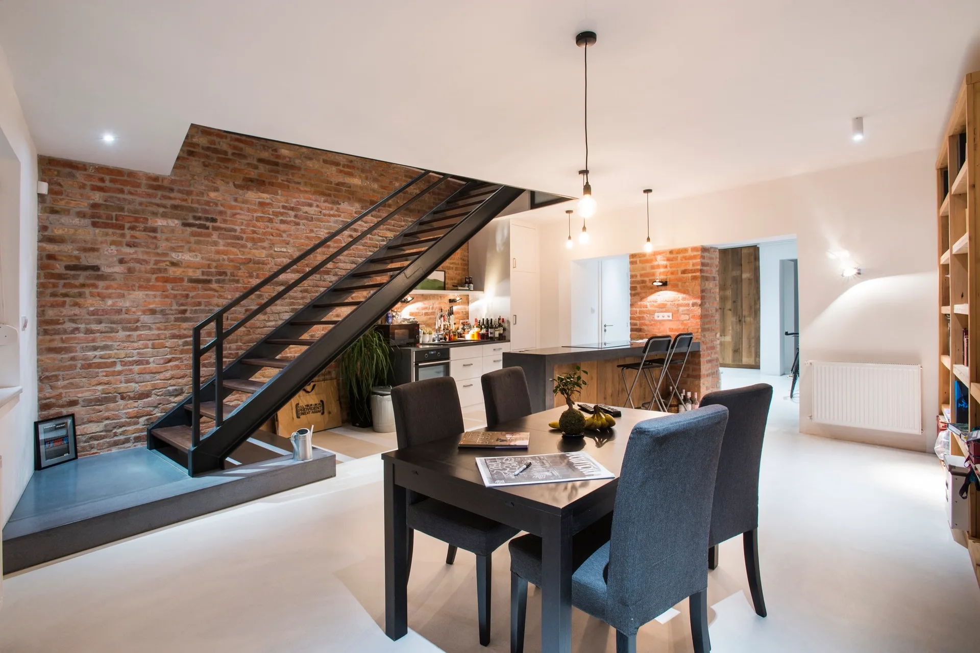 An open-concept dining room features a dark wood dining table with grey chairs, a black metal staircase, and an exposed brick wall.