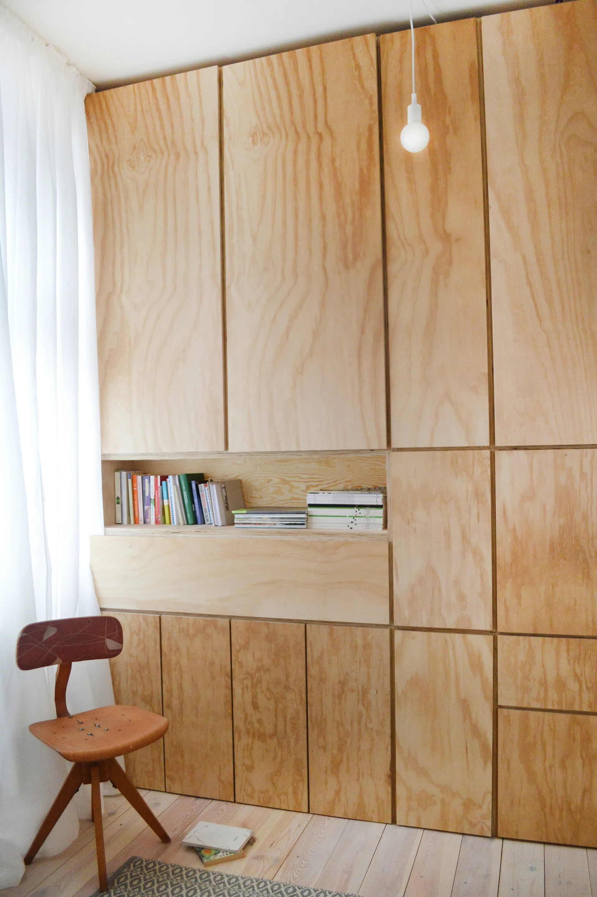 A floor-to-ceiling plywood storage unit with natural wood grain features a built-in horizontal niche for books, next to a white curtain and a vintage chair.