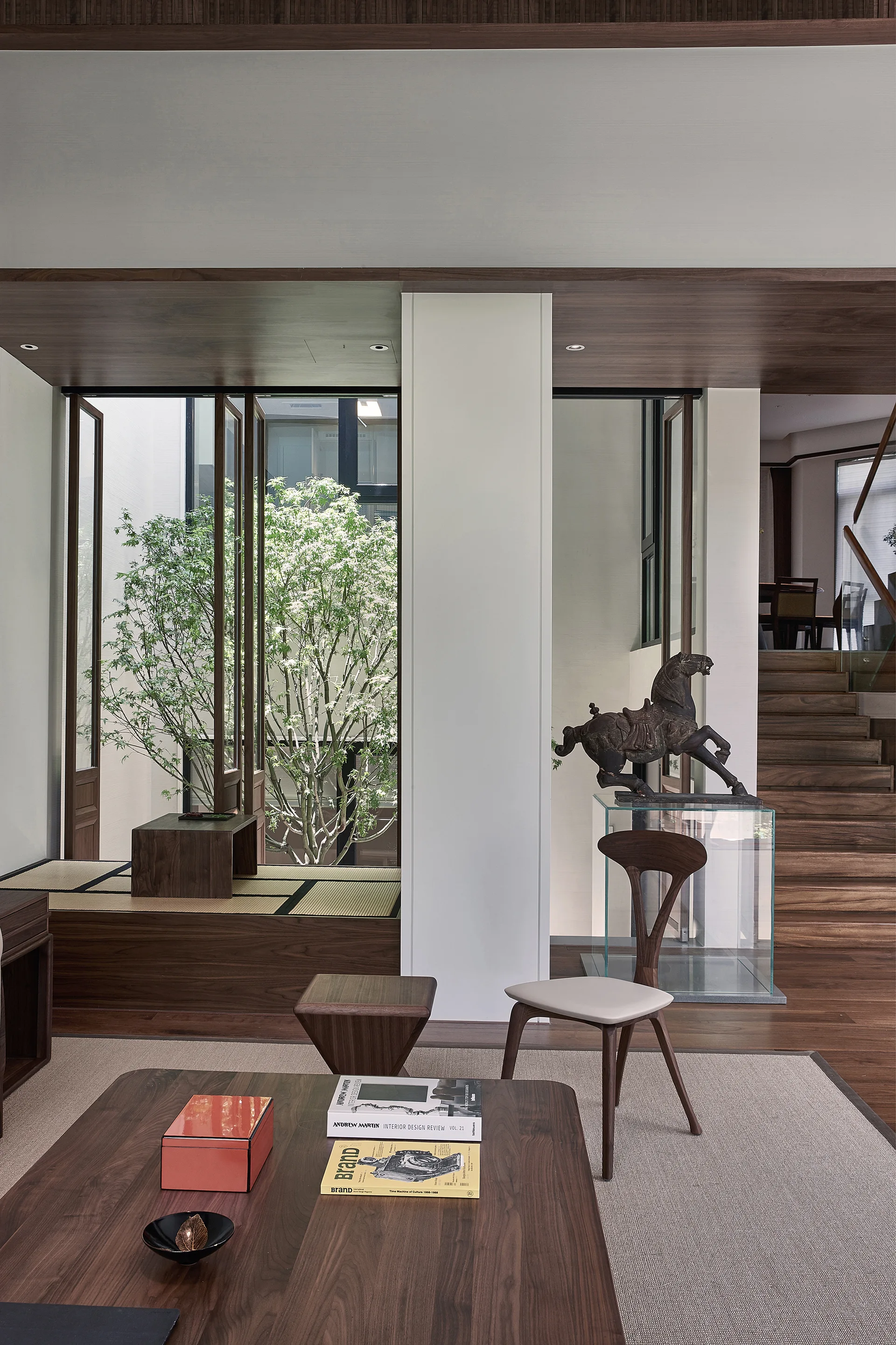 A minimalist living room featuring a warm wood coffee table, a raised tatami platform, and an interior courtyard with a tree.