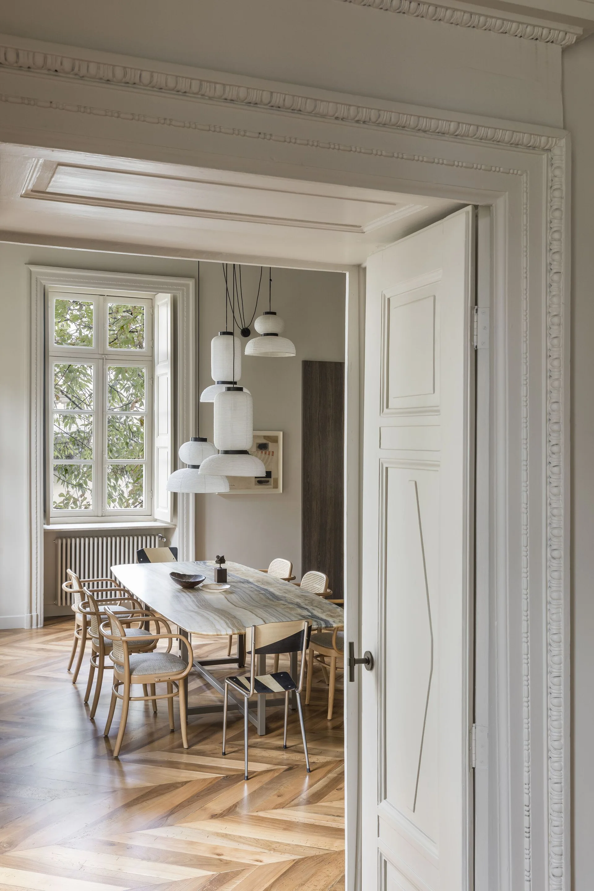 A dining room viewed through a doorway features a veined stone table, cane-backed chairs, and a cluster of white paper pendant lights over herringbone floors.