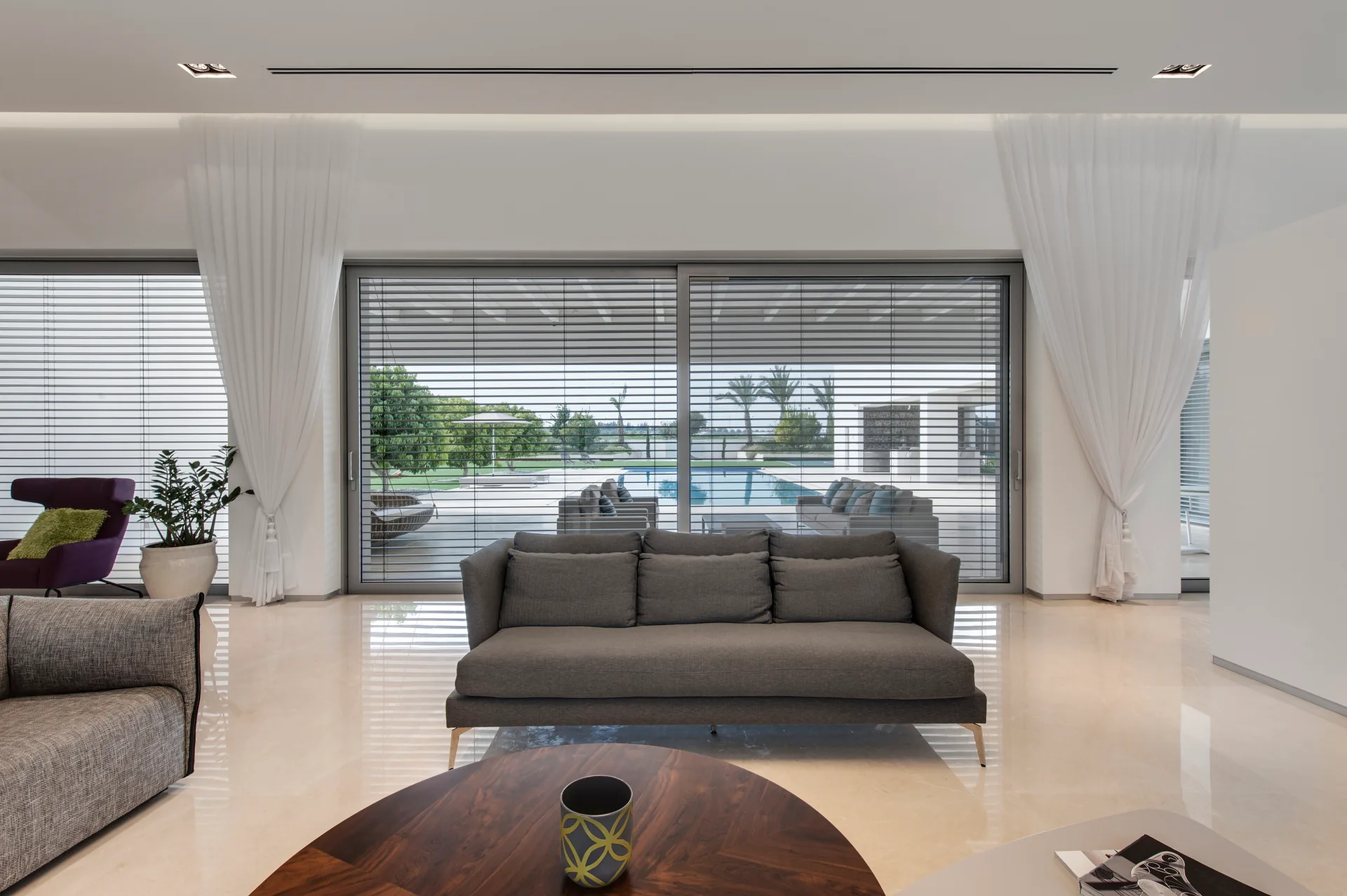 A modern living room features a dark grey sofa on polished light stone floors facing a large glass wall with horizontal blinds and a pool view.