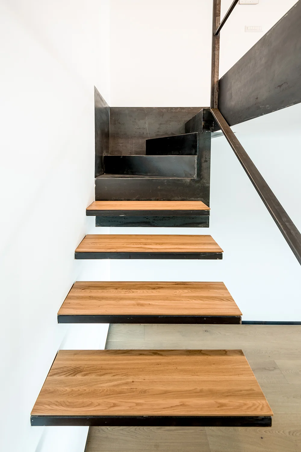 Minimalist floating staircase featuring natural wood treads mounted on a dark industrial metal frame against a plain white wall.