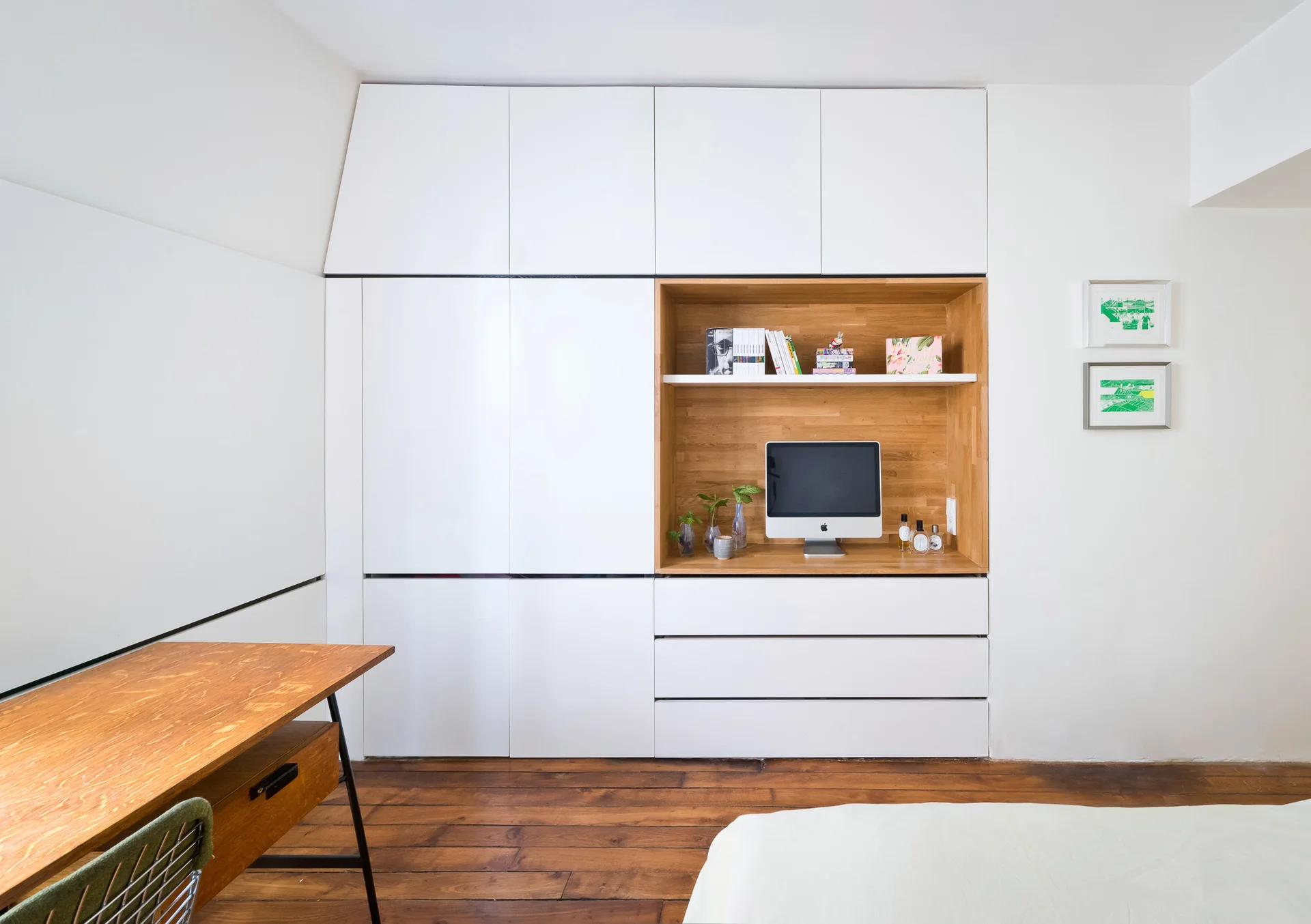 A white built-in wall cabinet features an integrated warm wood desk niche with a computer monitor, set against hardwood flooring.