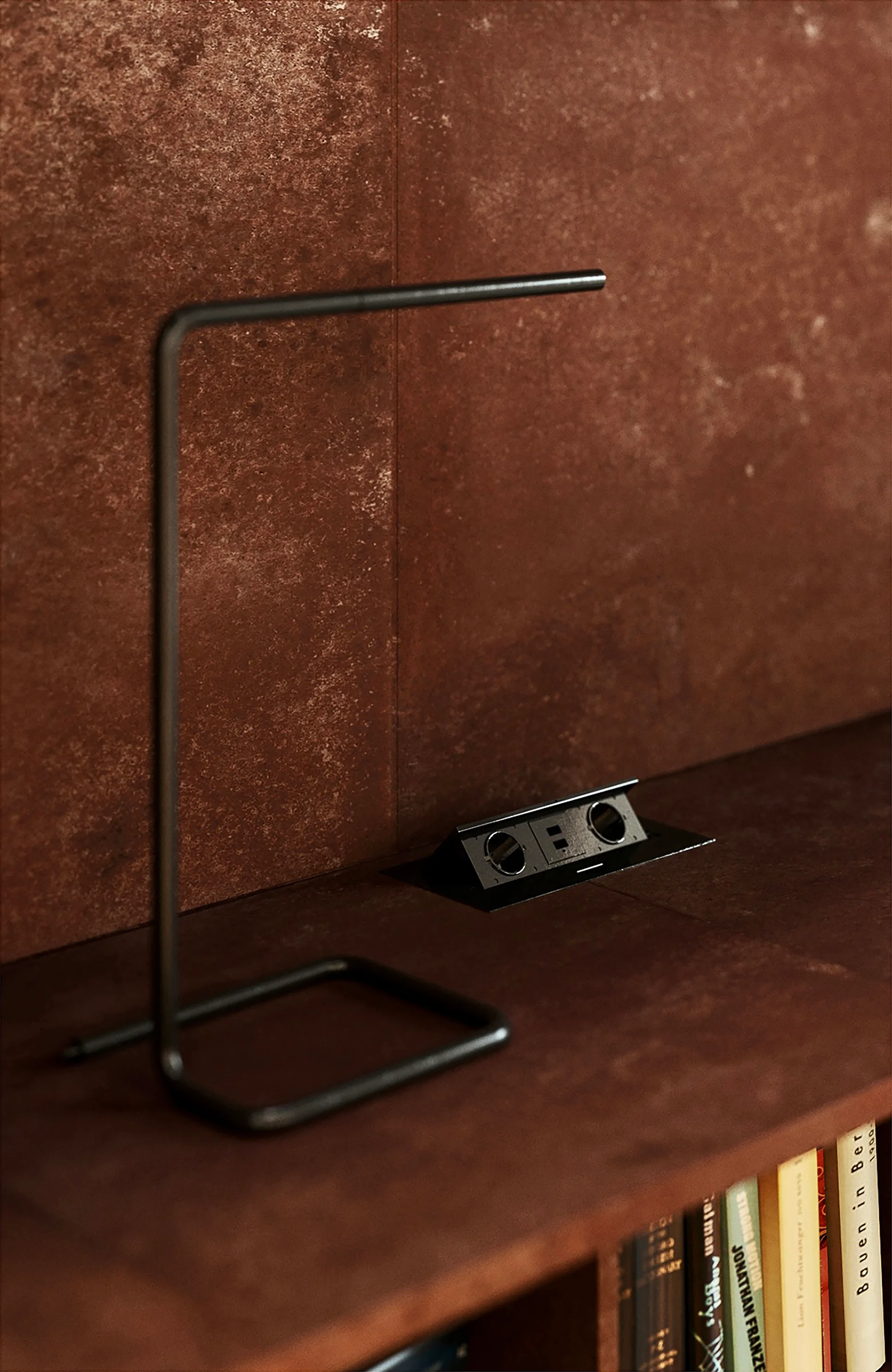A thin black metal desk lamp sits on a rustic brown textured shelf next to a recessed black electrical outlet with books visible below.
