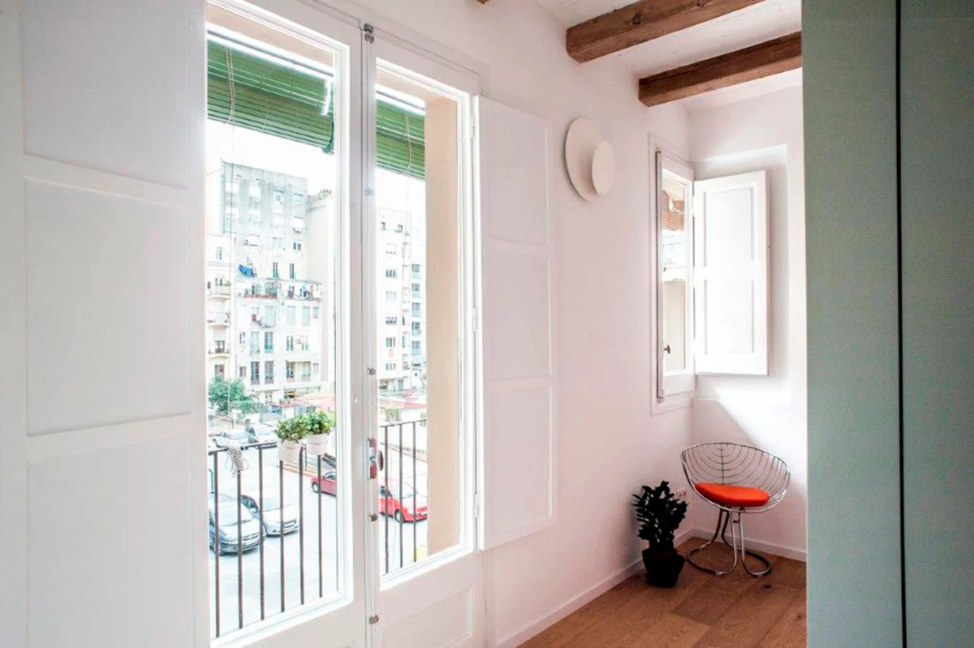 A bright room features white walls, exposed wooden ceiling beams, a wire chair with an orange cushion, and large glass doors leading to a balcony.