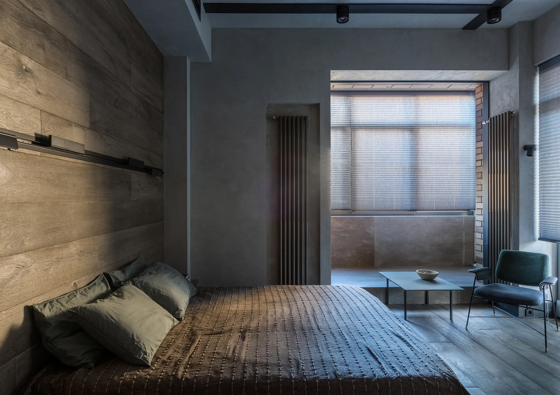 A modern bedroom features a bed with sage green pillows, a horizontal wood plank accent wall, concrete-textured gray walls, and a window nook.