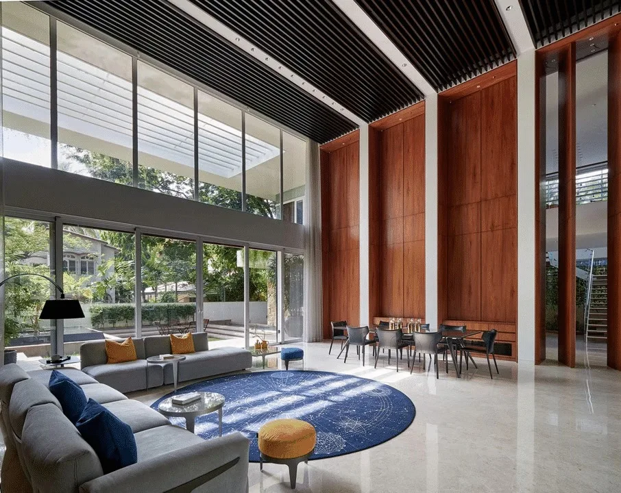 A spacious double-height contemporary living area featuring floor-to-ceiling windows, a large gray sectional sofa, and tall vertical wood wall panels.