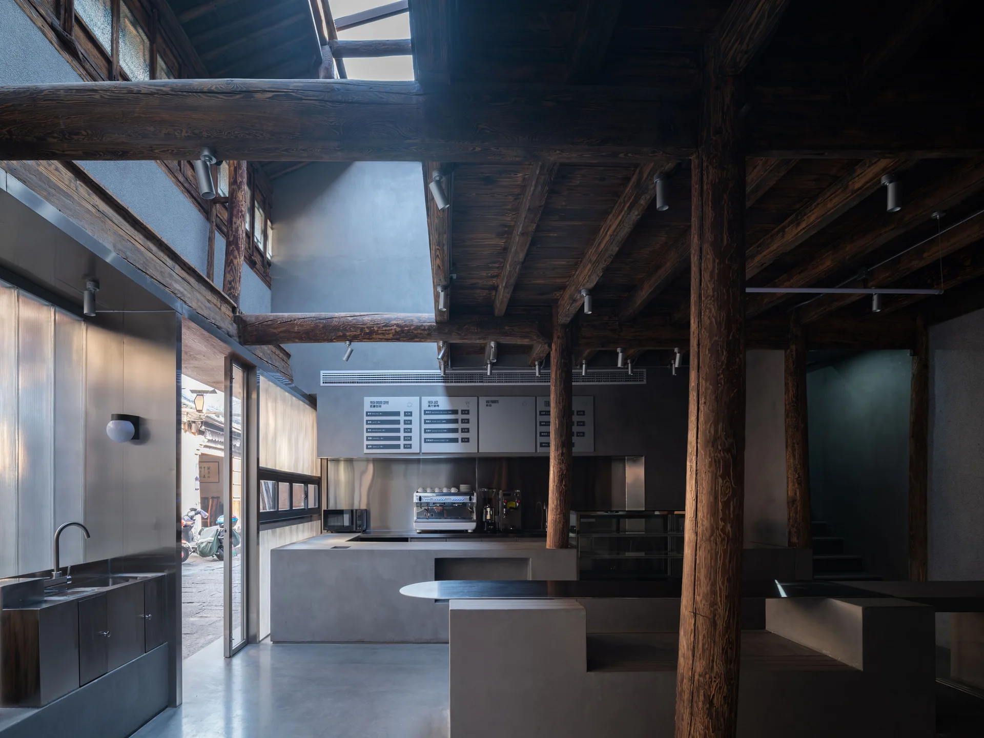 A modern cafe interior featuring monolithic concrete counters beneath a ceiling of heavy, dark rustic timber beams and a skylight.