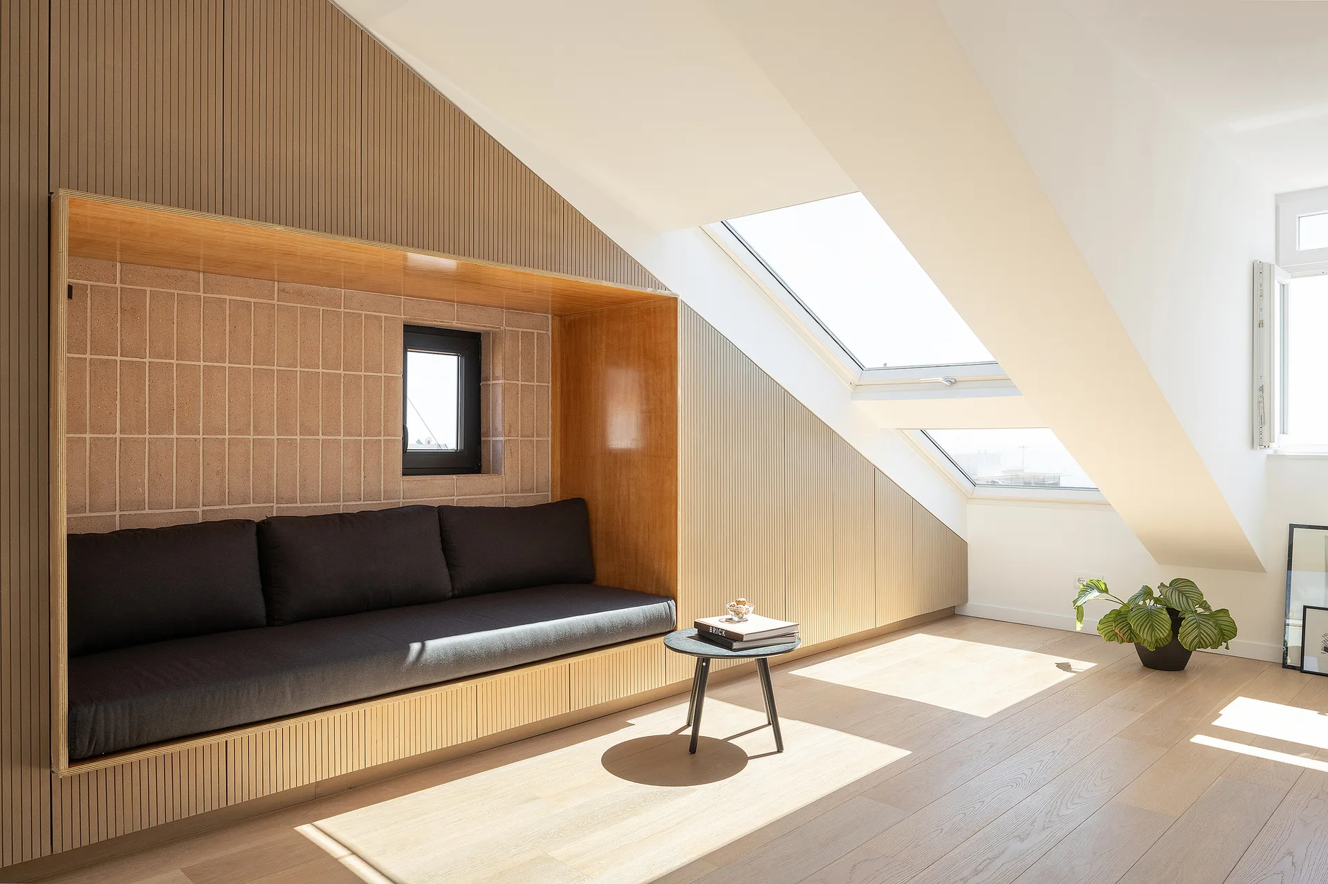 A minimalist attic living room features a built-in daybed niche lined with light wood and beige tile under a slanted ceiling with skylights.