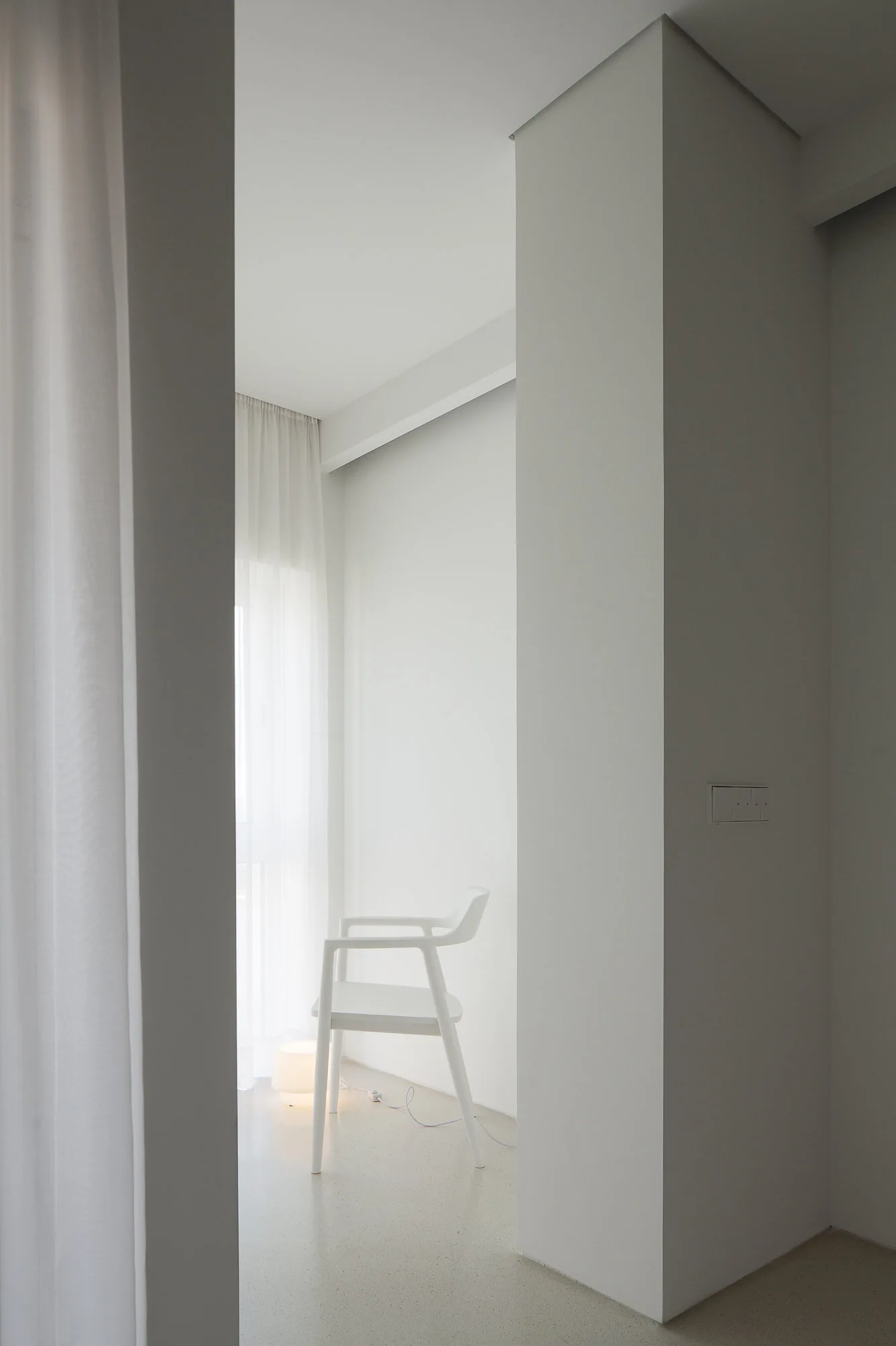 A single white modern armchair sits in a minimalist all-white room next to a window with sheer curtains and a low floor lamp.