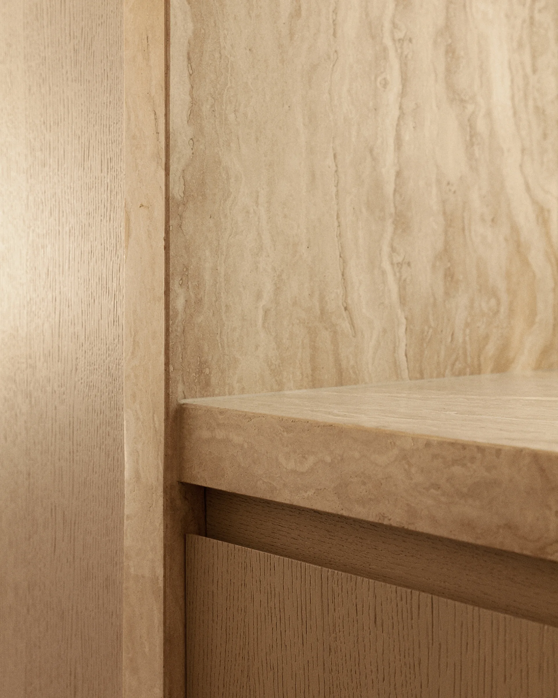 A close-up view of interior cabinetry shows a textured travertine stone shelf and wall panel meeting a light oak wood cabinet drawer and side panel.