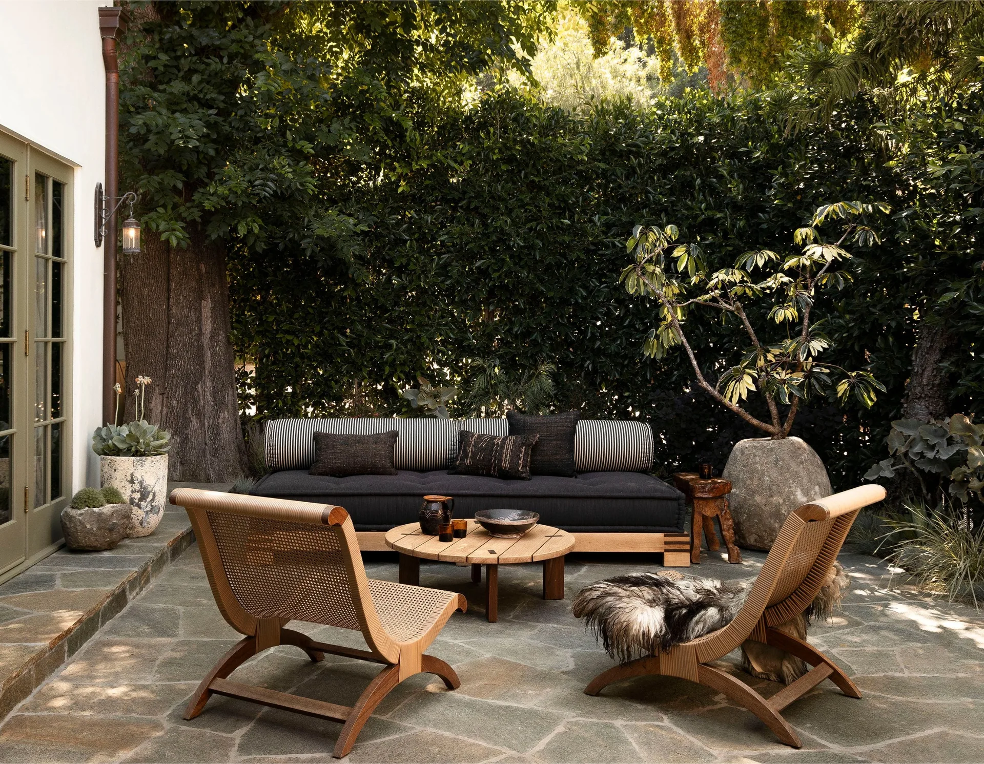 An outdoor flagstone patio features a low black sofa with a striped bolster, two curved wooden lounge chairs, and a round wood coffee table.