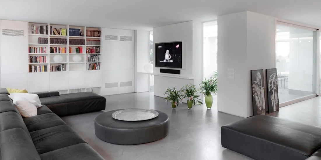 A minimalist living room features a dark leather sectional sofa, a large round ottoman, and extensive white built-in bookcases on a grey concrete floor.