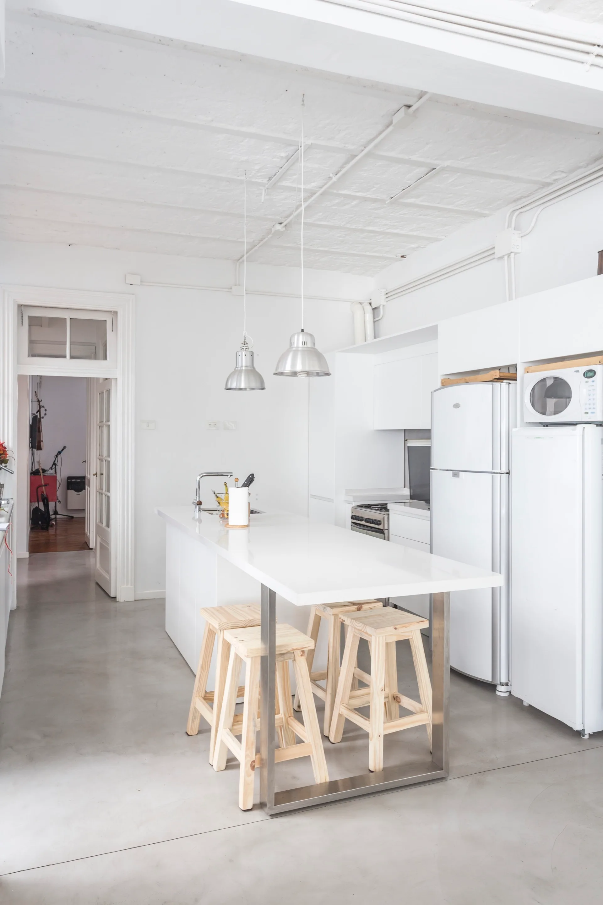 A minimalist white kitchen featuring a central island with four light wood stools, concrete flooring, and metallic pendant lights.