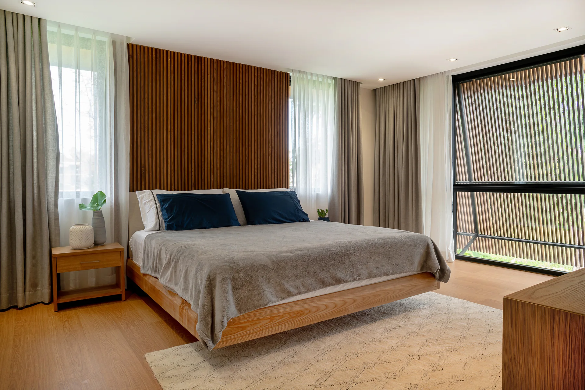 A modern bedroom features a wooden platform bed with blue pillows positioned against a tall vertical wood slat accent wall framed by grey curtains.