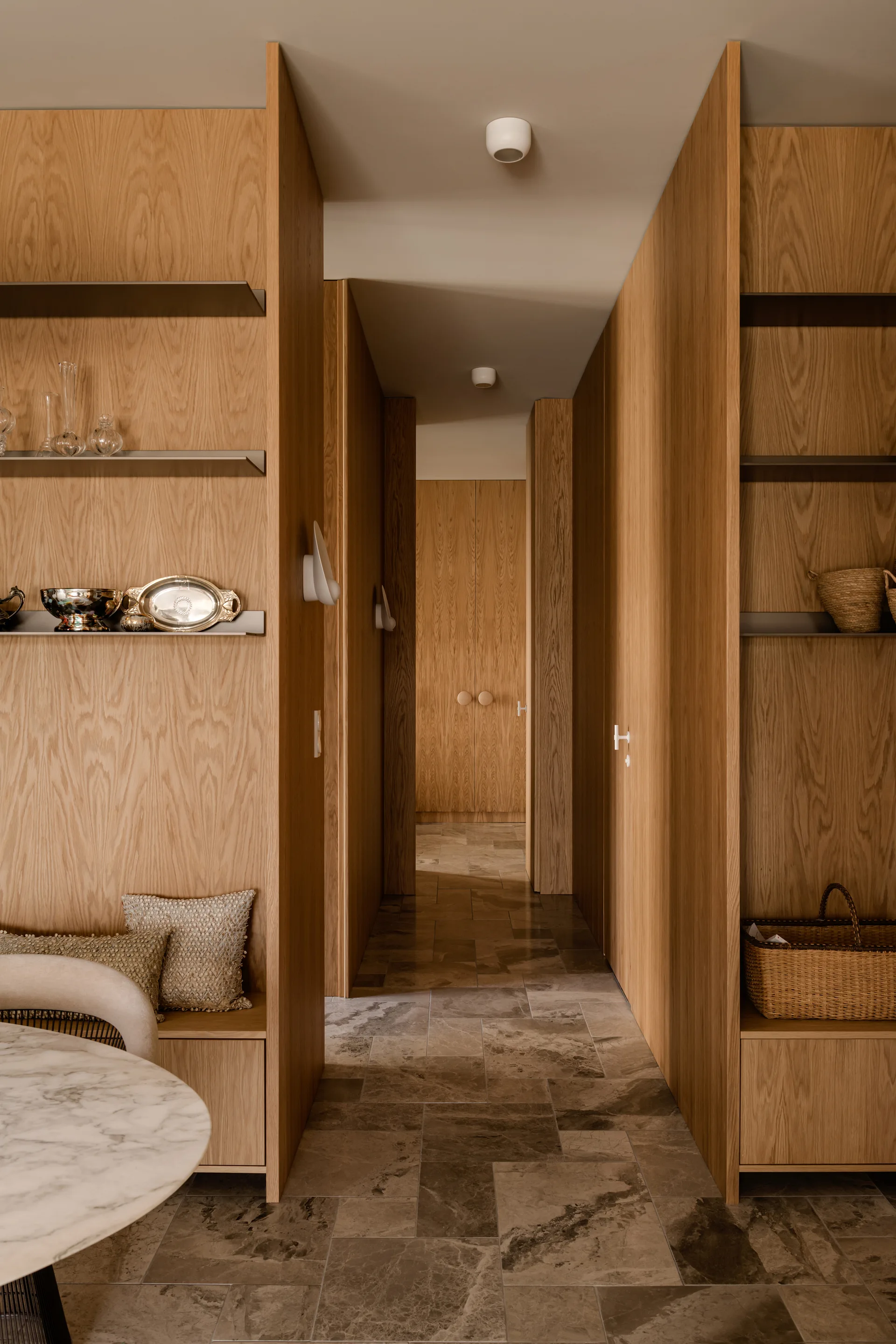 A hallway featuring floor-to-ceiling oak veneer cabinetry, integrated display shelves, and a textured grey stone tile floor.