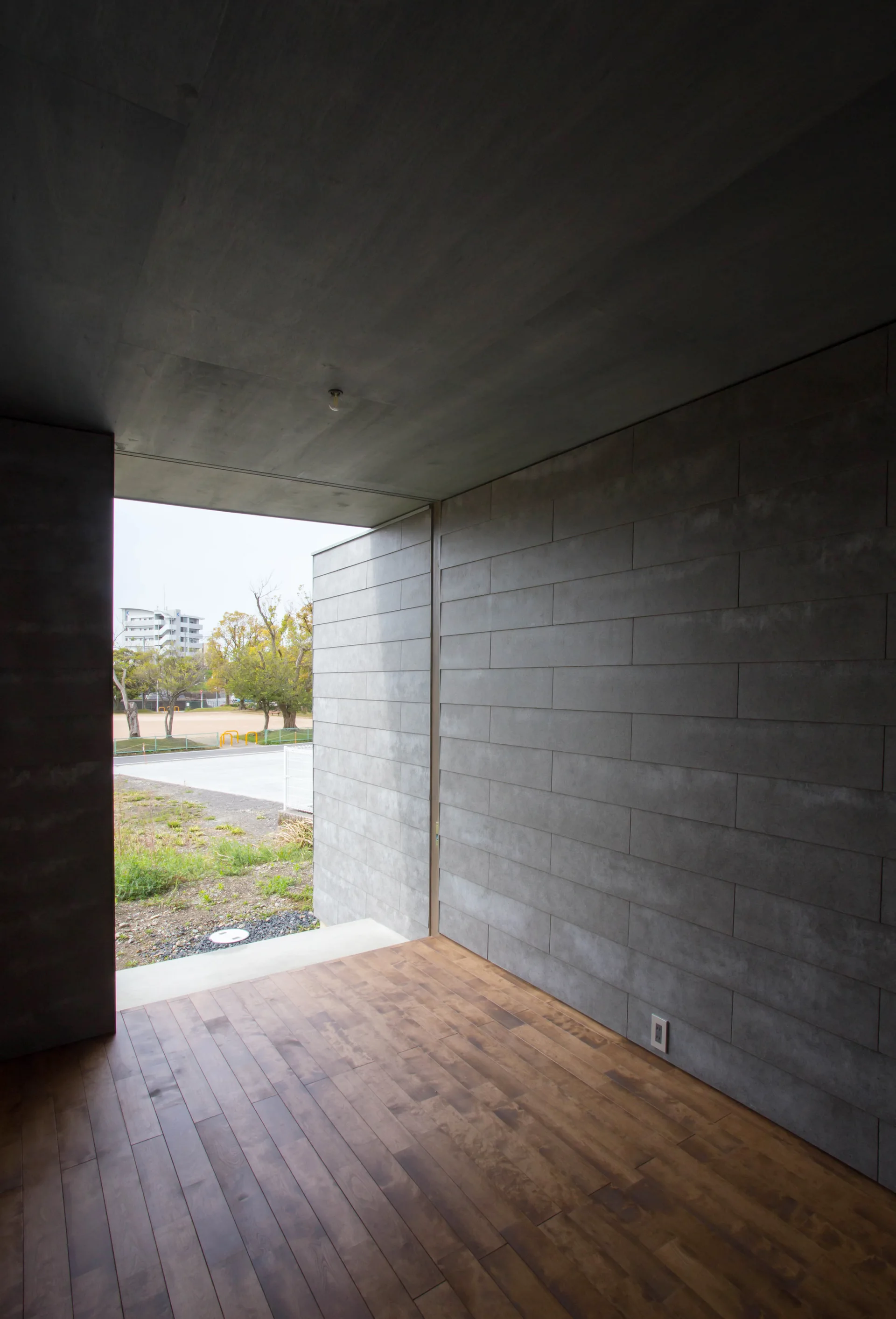 A minimalist room features grey tiled walls, wood plank flooring, and a large open threshold leading to an outdoor landscape.