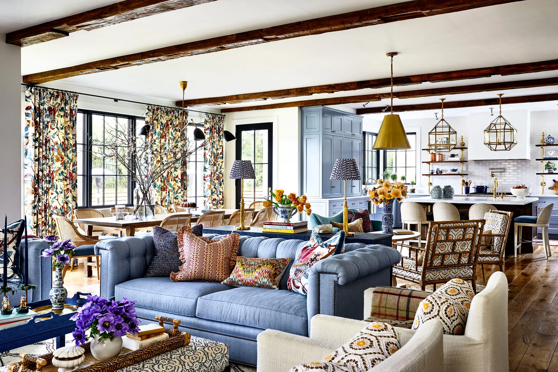 A transitional open-concept living room featuring a blue tufted sofa, patterned drapes, dark ceiling beams, and a blue kitchen in the background.